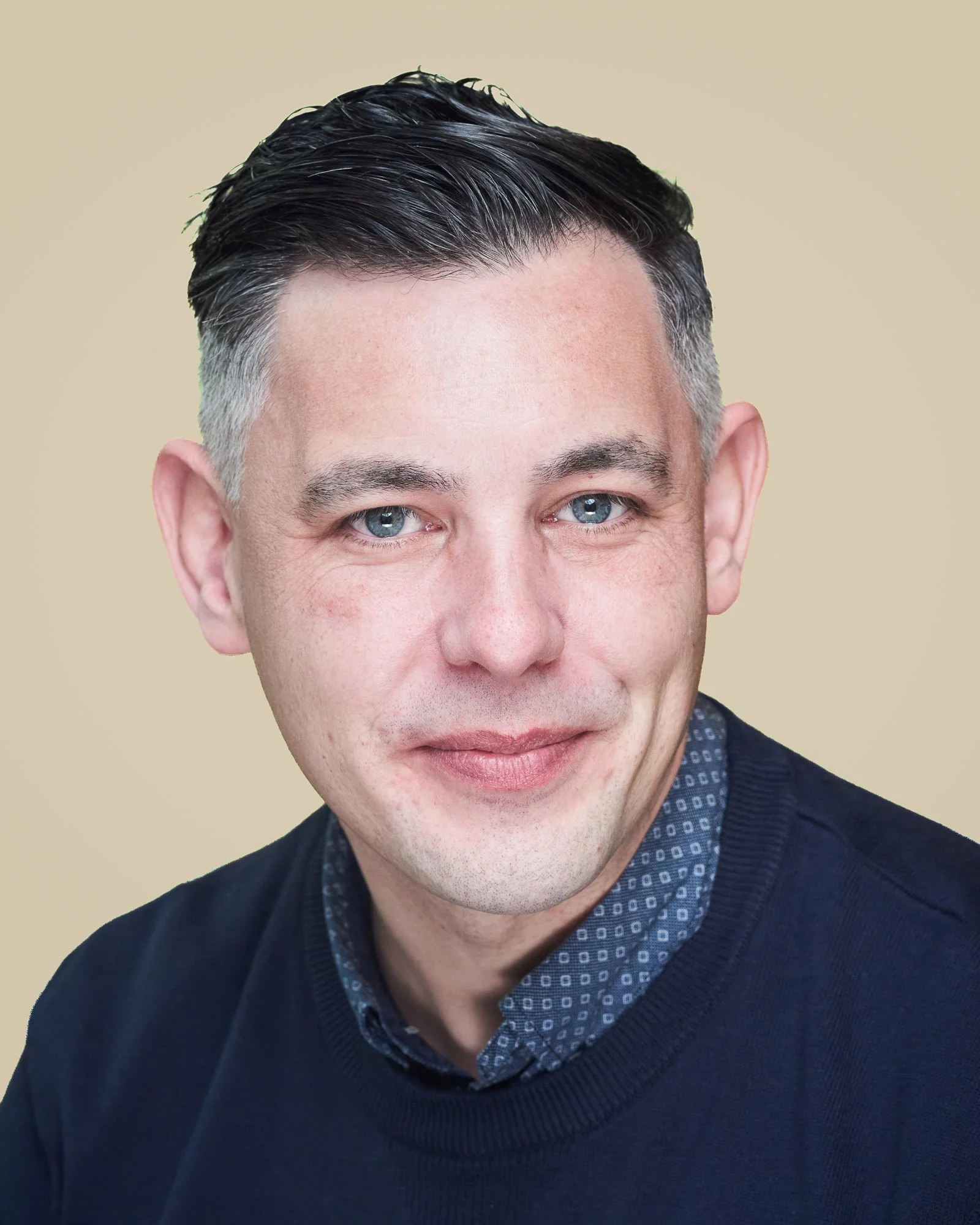 Close-up of a man's face with blue eyes, dark hair, and wearing a dark sweater and patterned shirt, against a beige background.