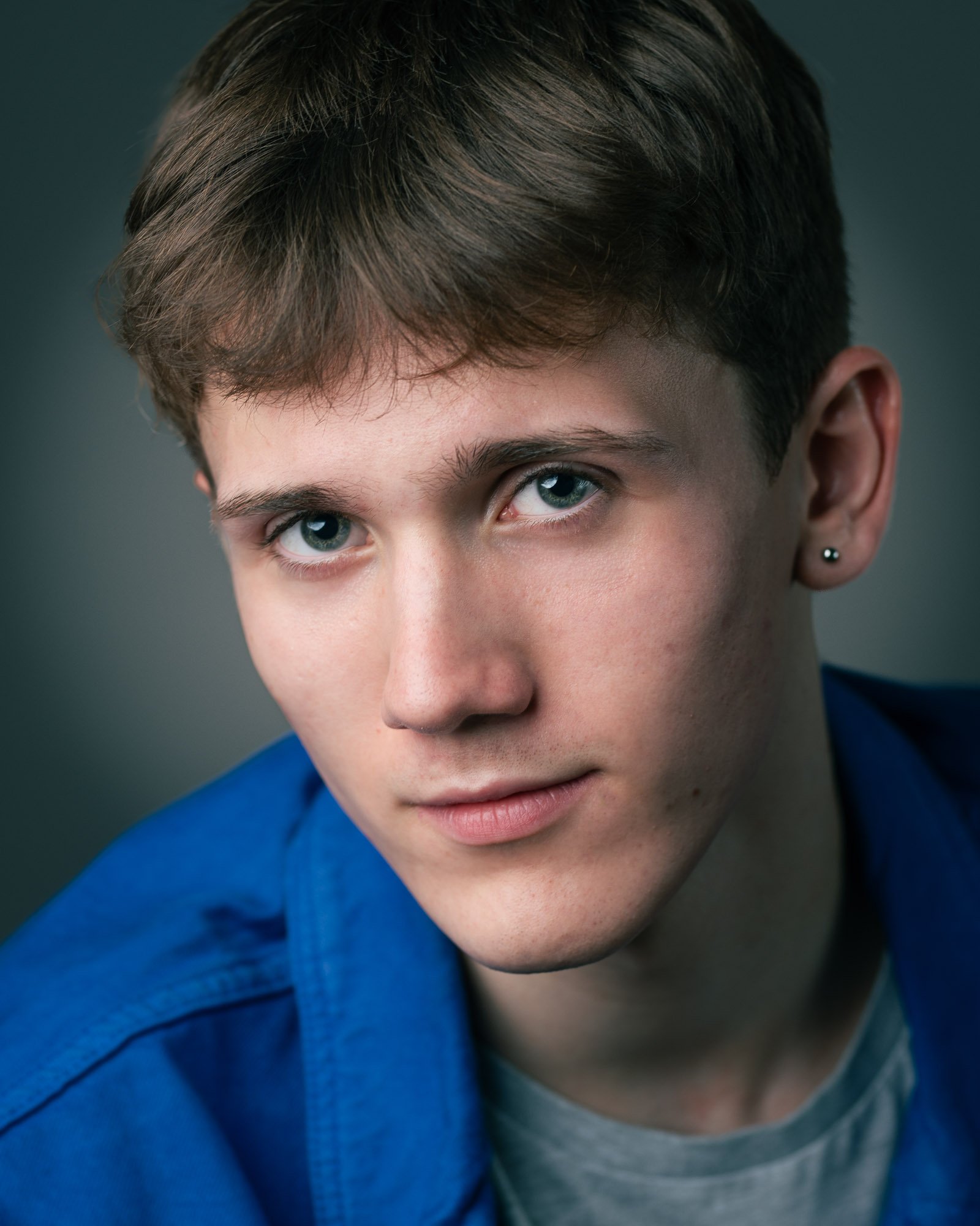 Close-up of a young person's face with short brown hair, blue eyes, earrings, wearing a blue jacket and gray shirt.