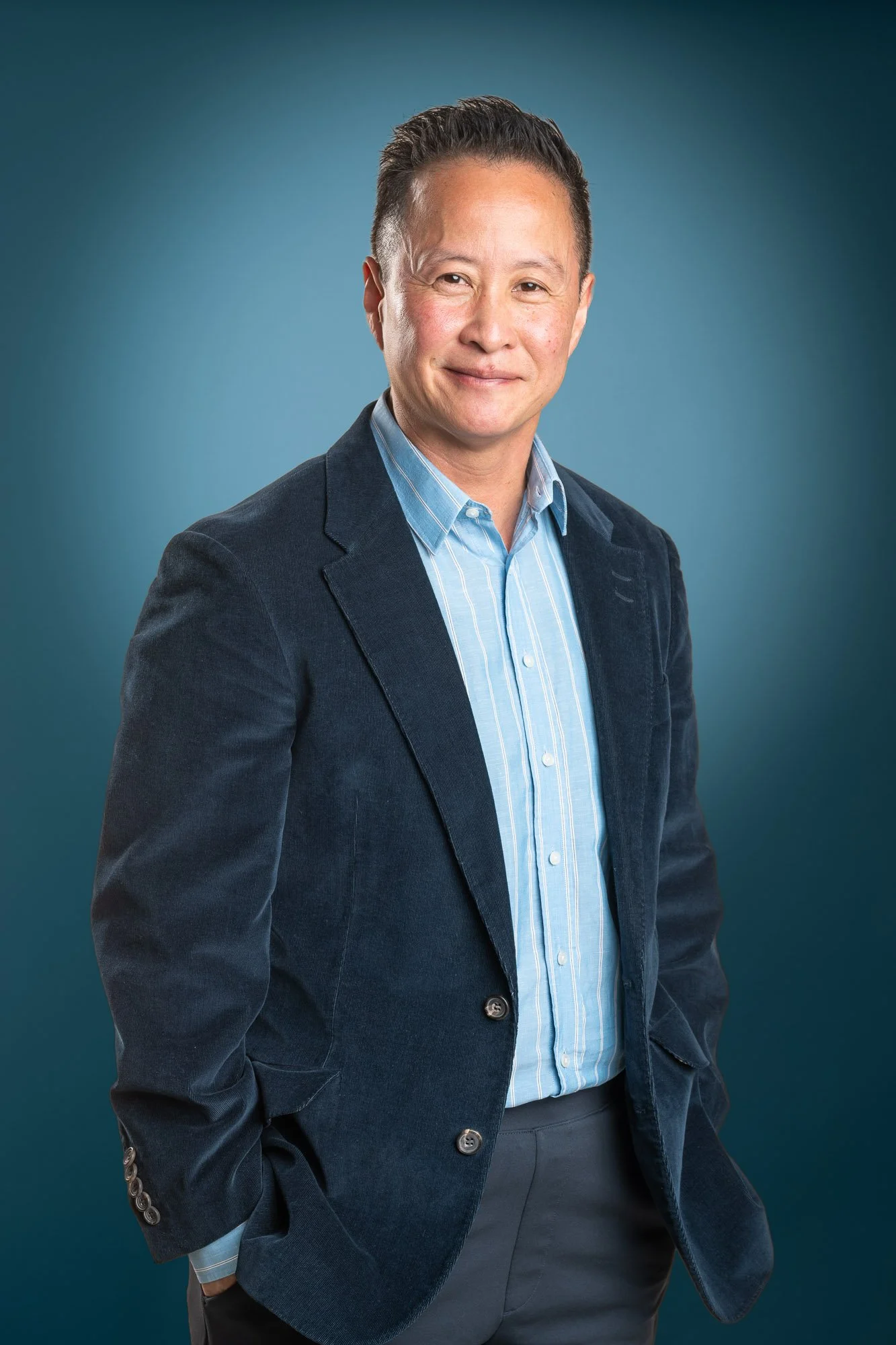 A professional man with short dark hair, wearing a blue striped shirt and a navy blazer, posing against a blue background.