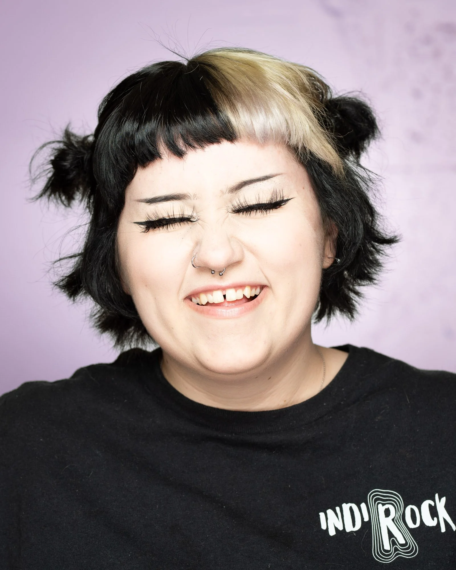 A woman with black and blonde hair, smiling with teeth showing, wearing a black T-shirt with 'IND'ROCK' printed on it, against a light purple background.