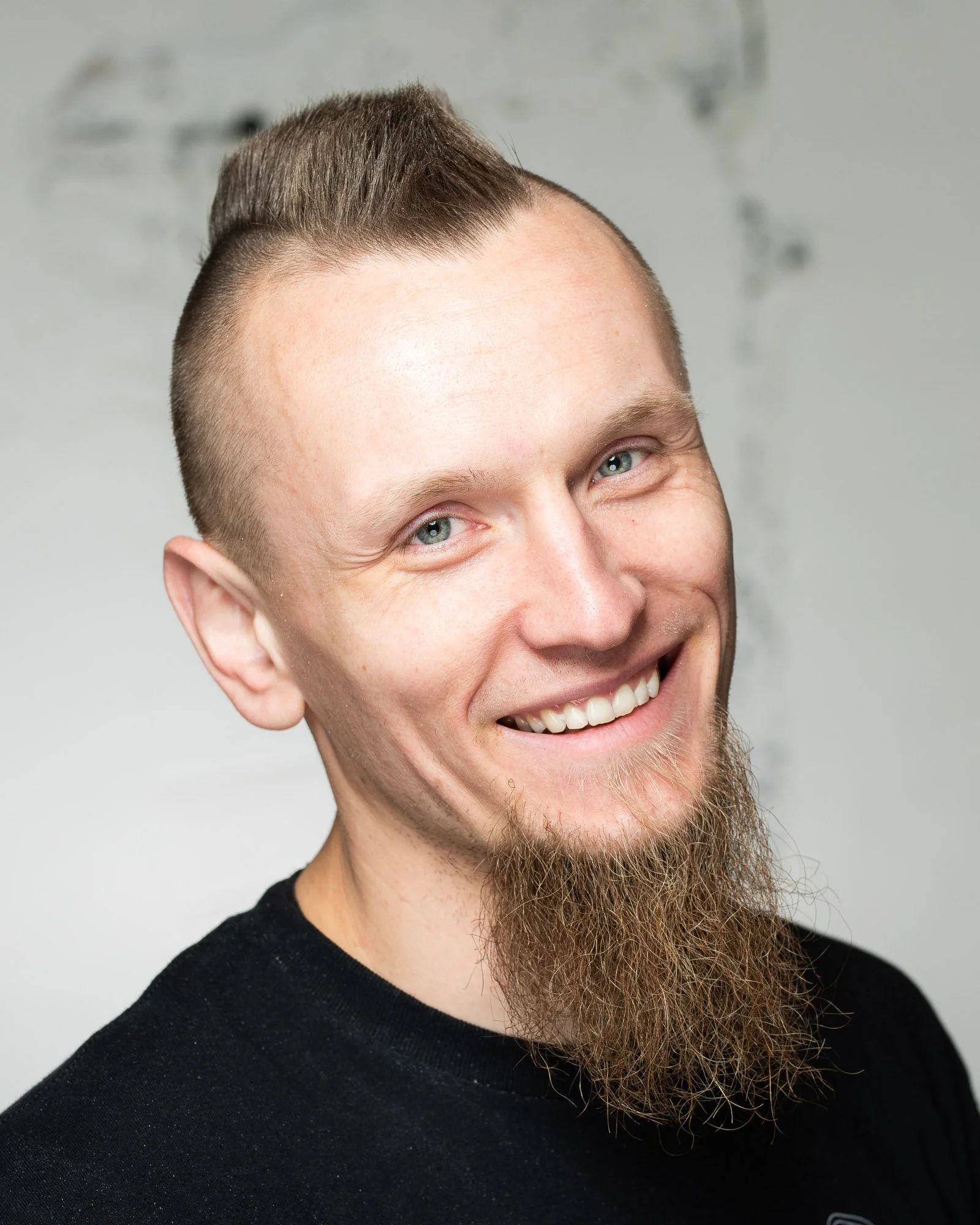 Close-up of a smiling man with short styled hair, light skin, blue eyes, and a long beard wearing a black shirt, against a light background.