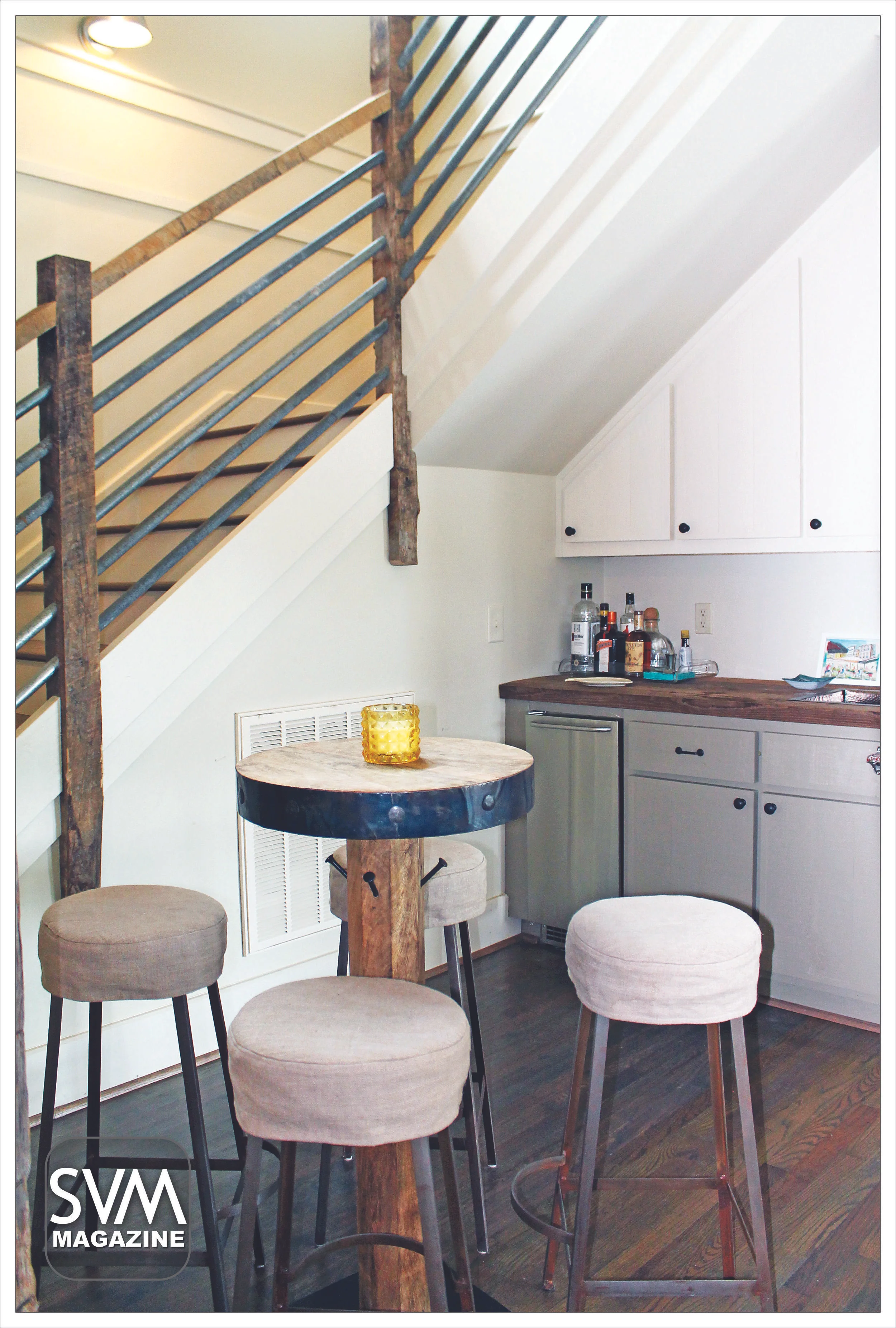 The stair railing, made with reclaimed wood by Tim Gregory, leads you to the newly built, second floor of the home. The high ceilings allow for a wet bar and seating area under the stairs. The wooden legged bar stools and reclaimed wood counter top add rustic charm to this space.