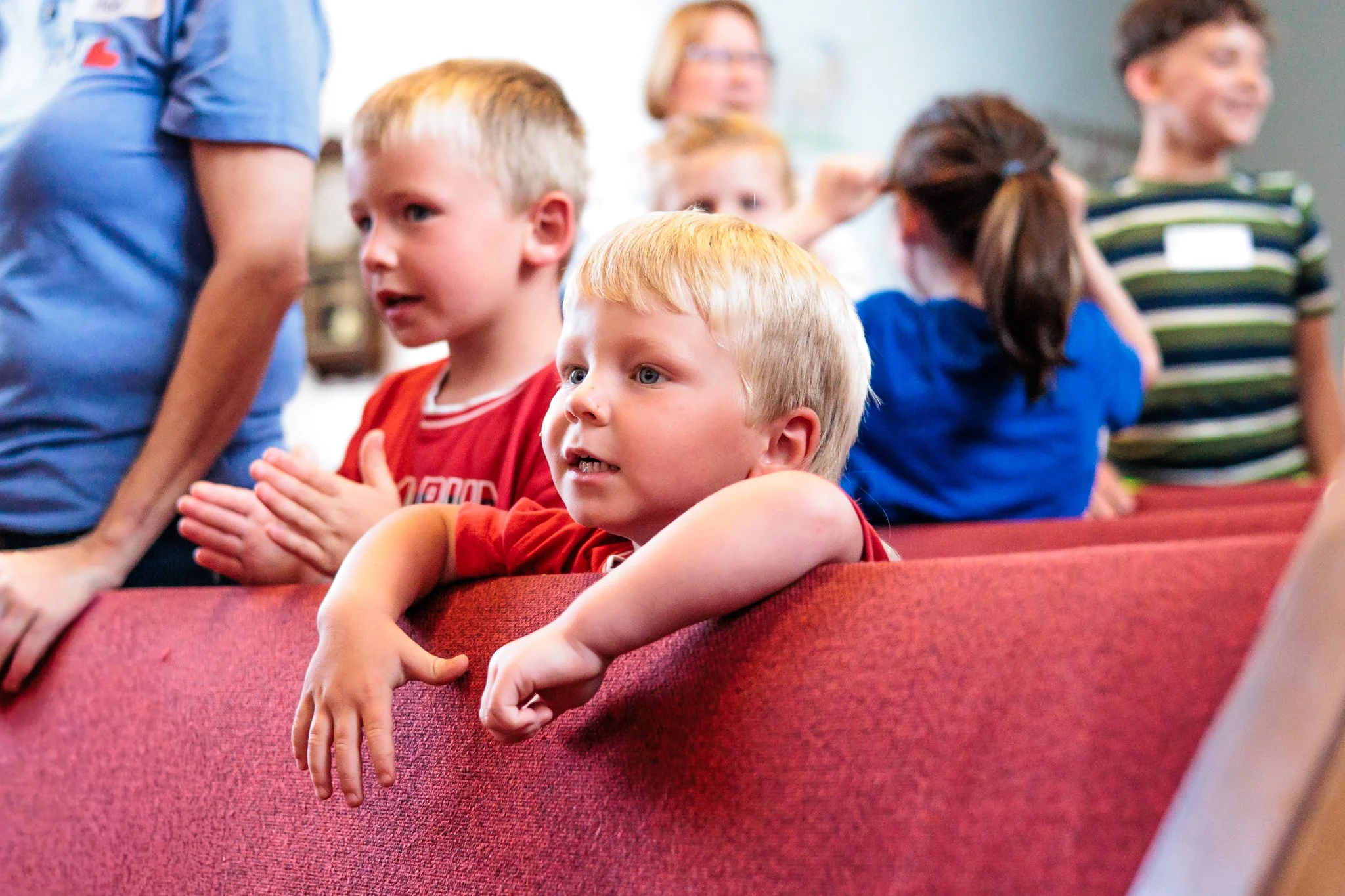 Children sitting on red theater seats, attentively watching a performance or presentation in an indoor venue.