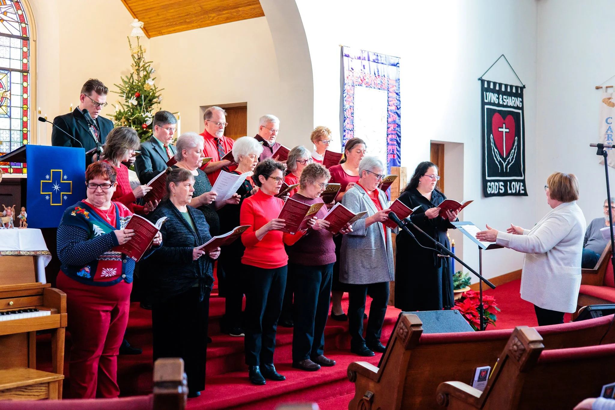 A choir of people singing and reading sheet music in a church with Christmas decorations, including a decorated Christmas tree and poinsettias, during a Christmas service.