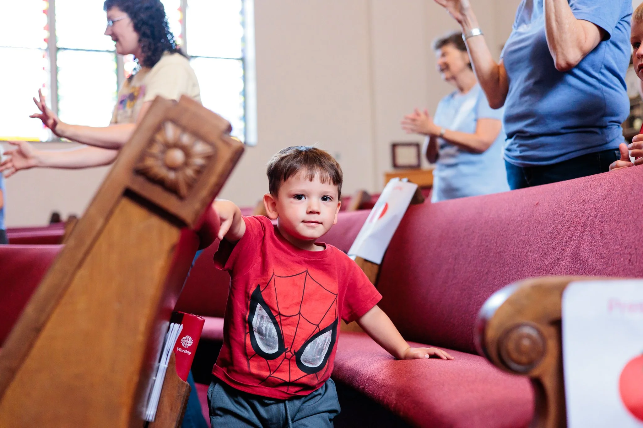 A young boy in a red Spider-Man t-shirt is kneeling on the church pews, looking directly at the camera with a serious expression. Several adults are standing behind him, with some raising their hands in prayer or worship during a church service.