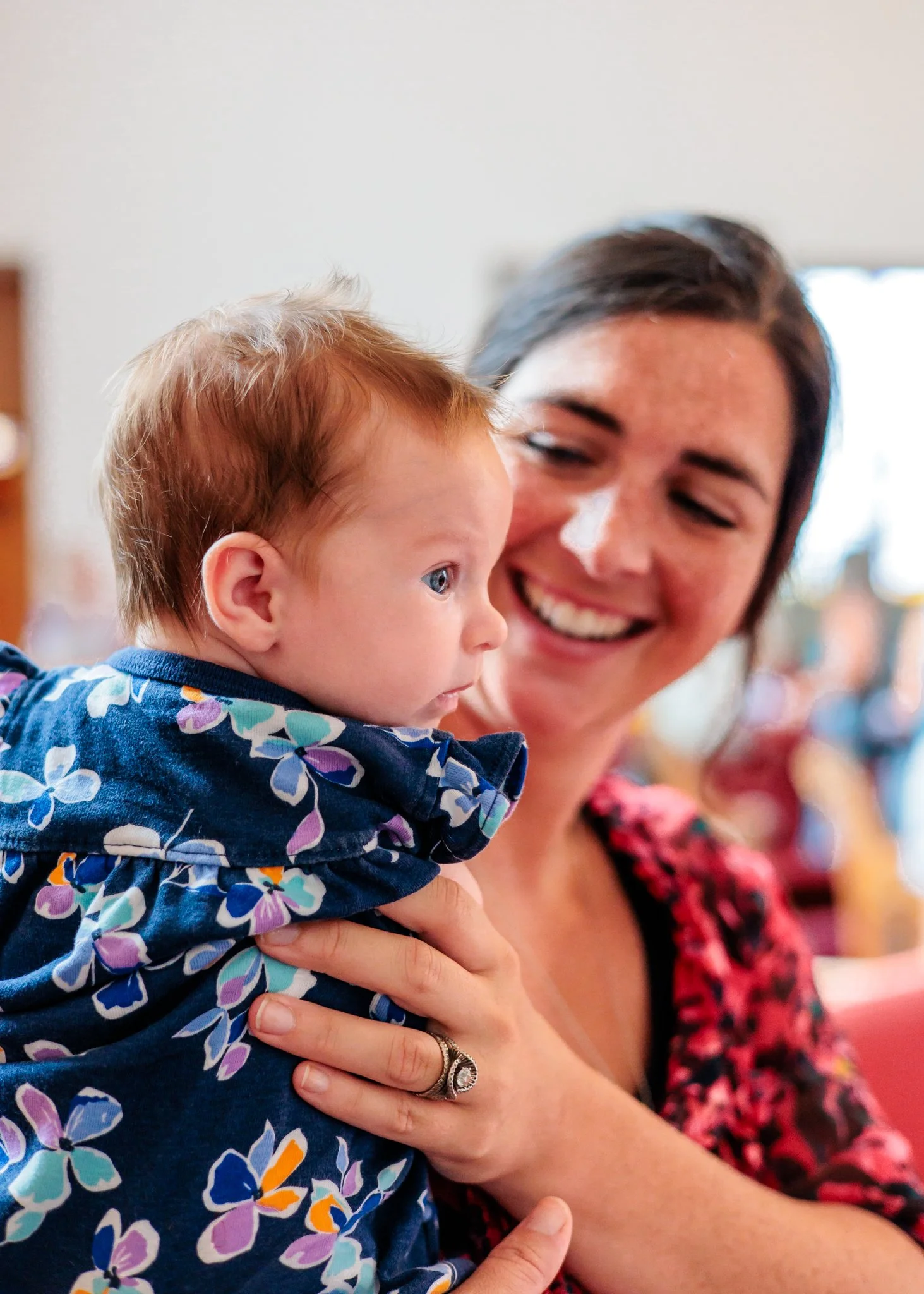 A woman holding a young red-haired boy in a blue floral shirt, smiling and looking at him, with another woman blurred in the background.