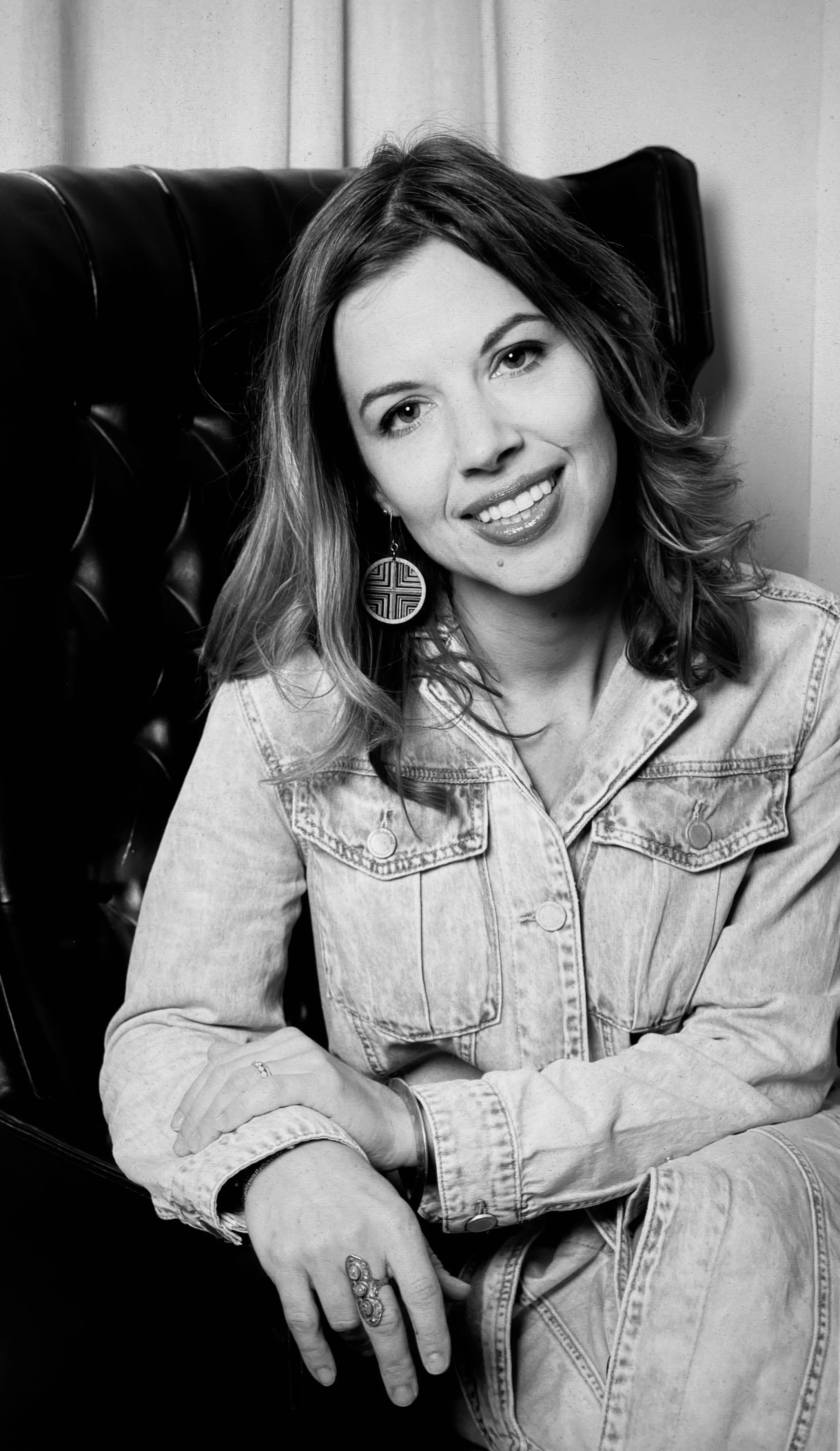 A black and white photo of a woman with shoulder-length wavy hair, wearing earrings and a denim jacket, sitting with crossed arms and a smile, in front of a black leather cushioned seat.
