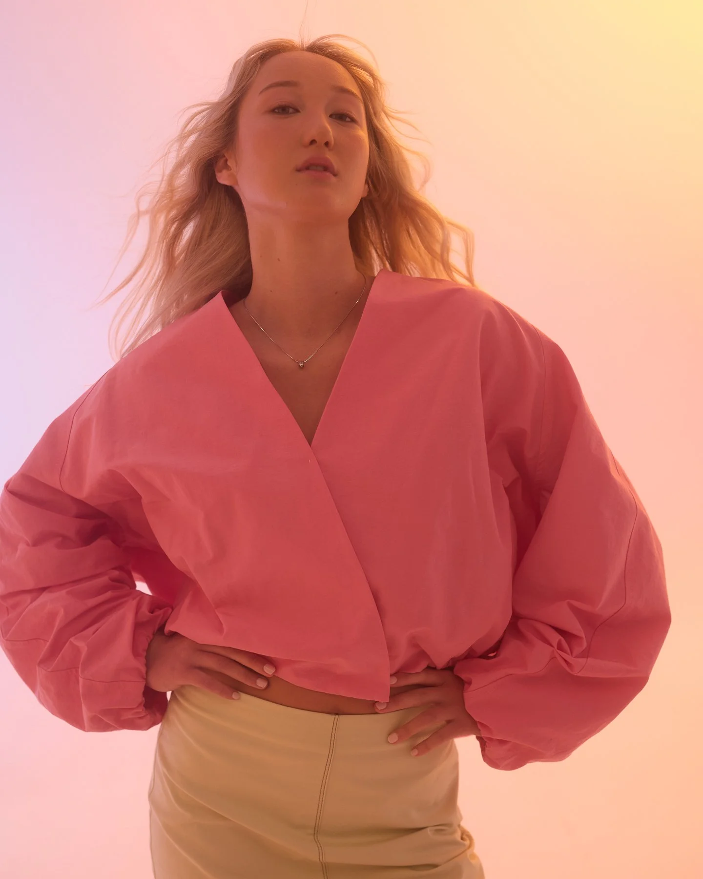 A woman with blonde hair wearing a pink top and beige skirt, standing against a warm-colored background.