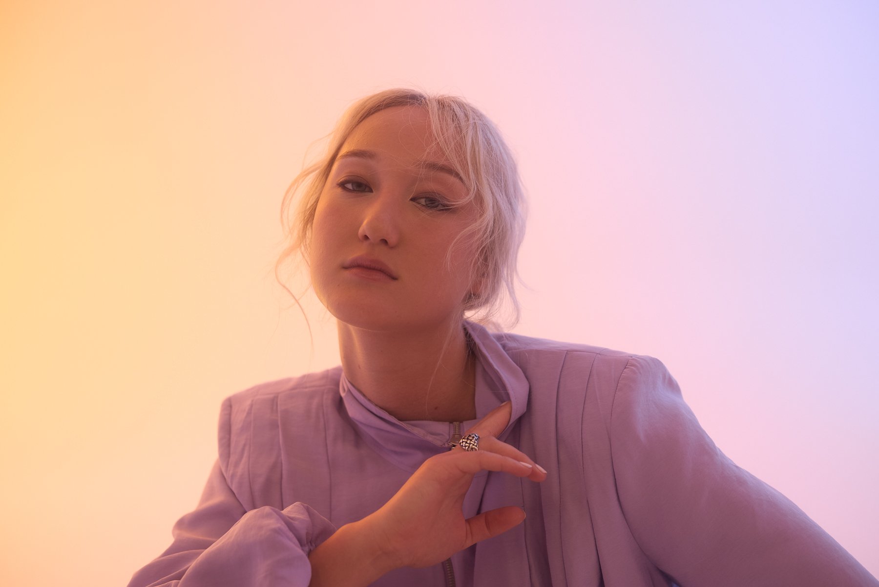 A young woman with blonde hair and a neutral expression, wearing a light purple jacket, is posing against a pastel background with gradient colors of pink, purple, and yellow.