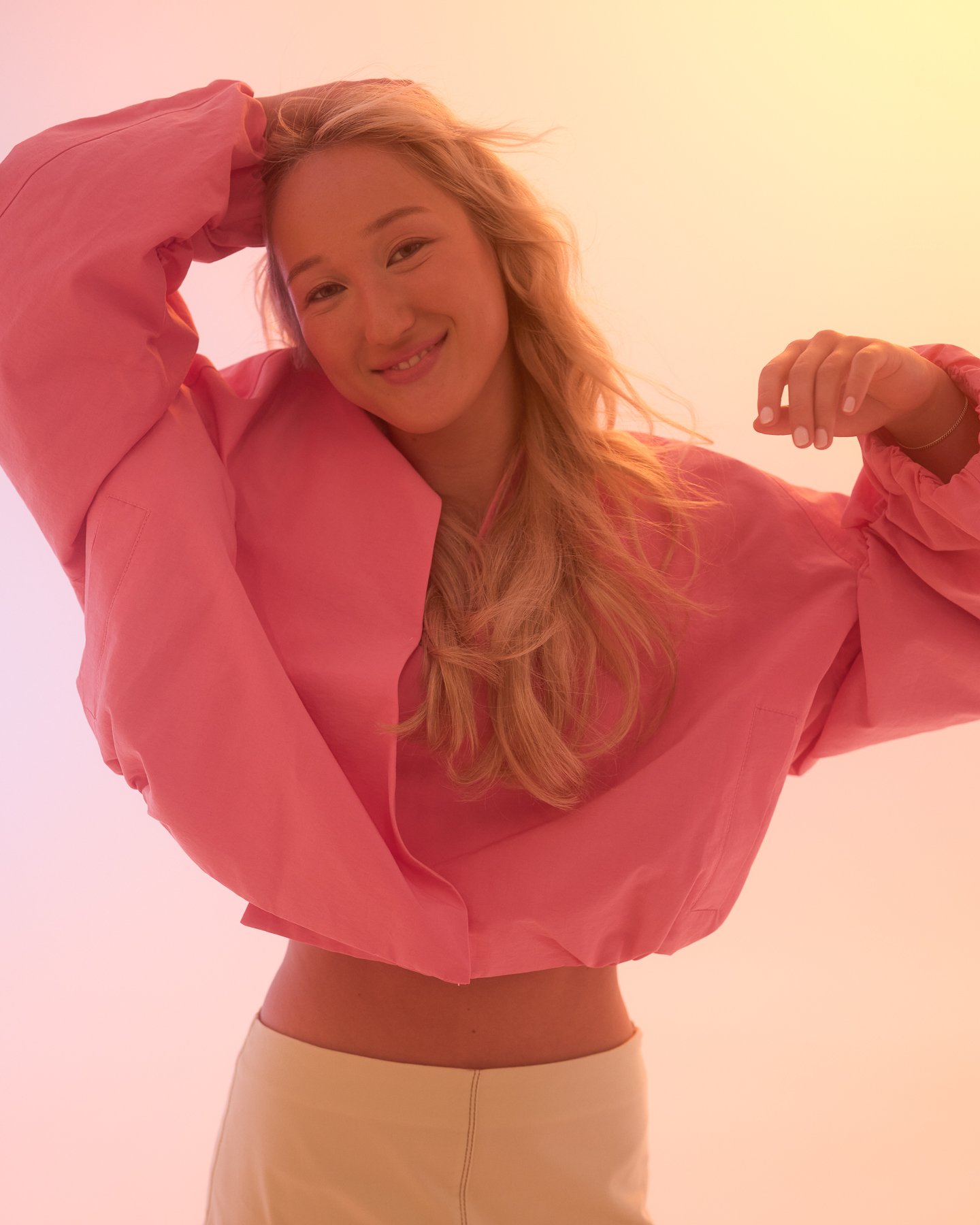 A young woman with blonde hair and light skin, wearing a pink jacket and beige pants, smiling and posing with one hand behind her head and the other bent at the elbow against a light pink background.