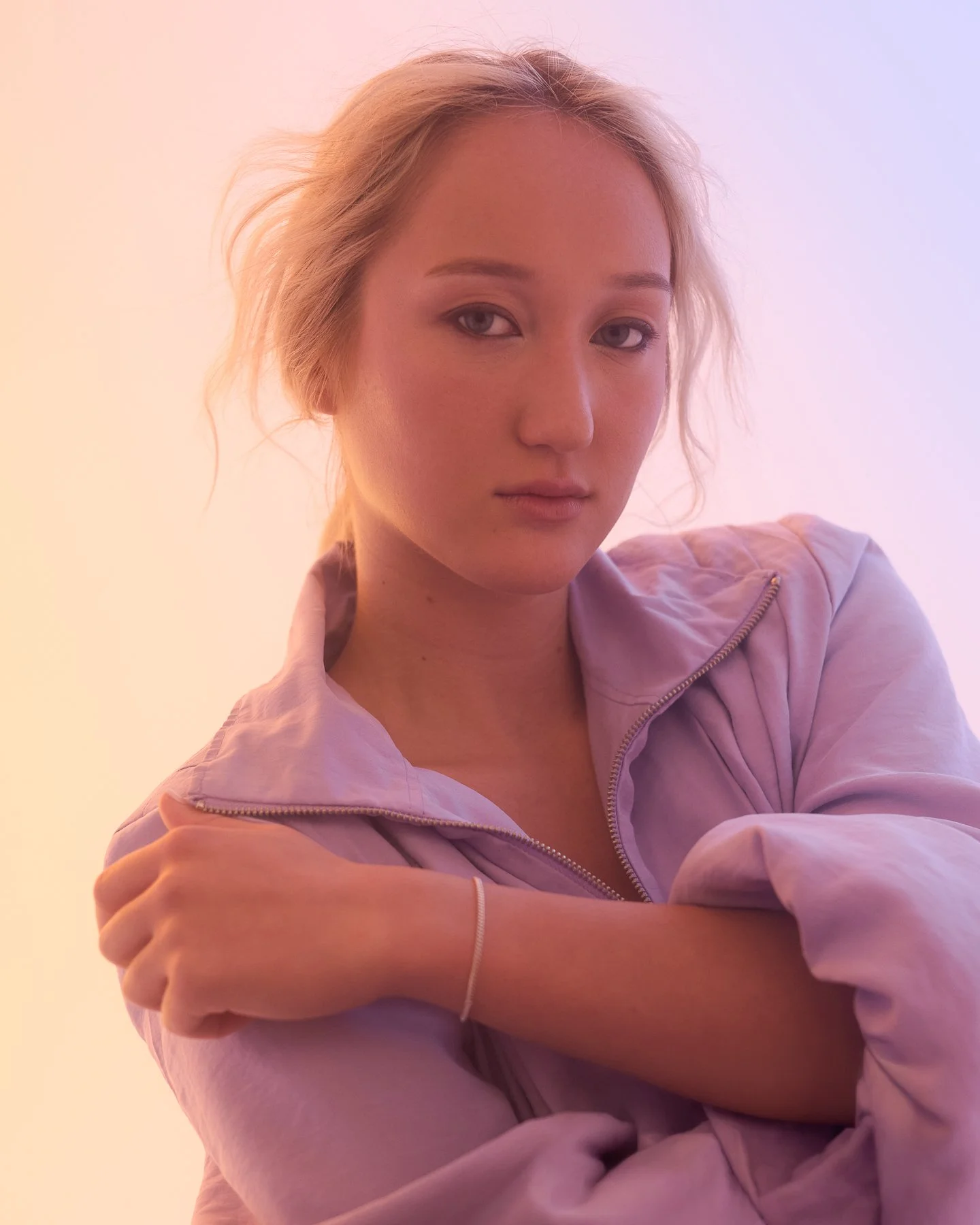 A woman with fair skin and blonde wavy hair, wearing a light-colored zip-up jacket, looking at the camera with a neutral expression, set against a soft pink and purple background.