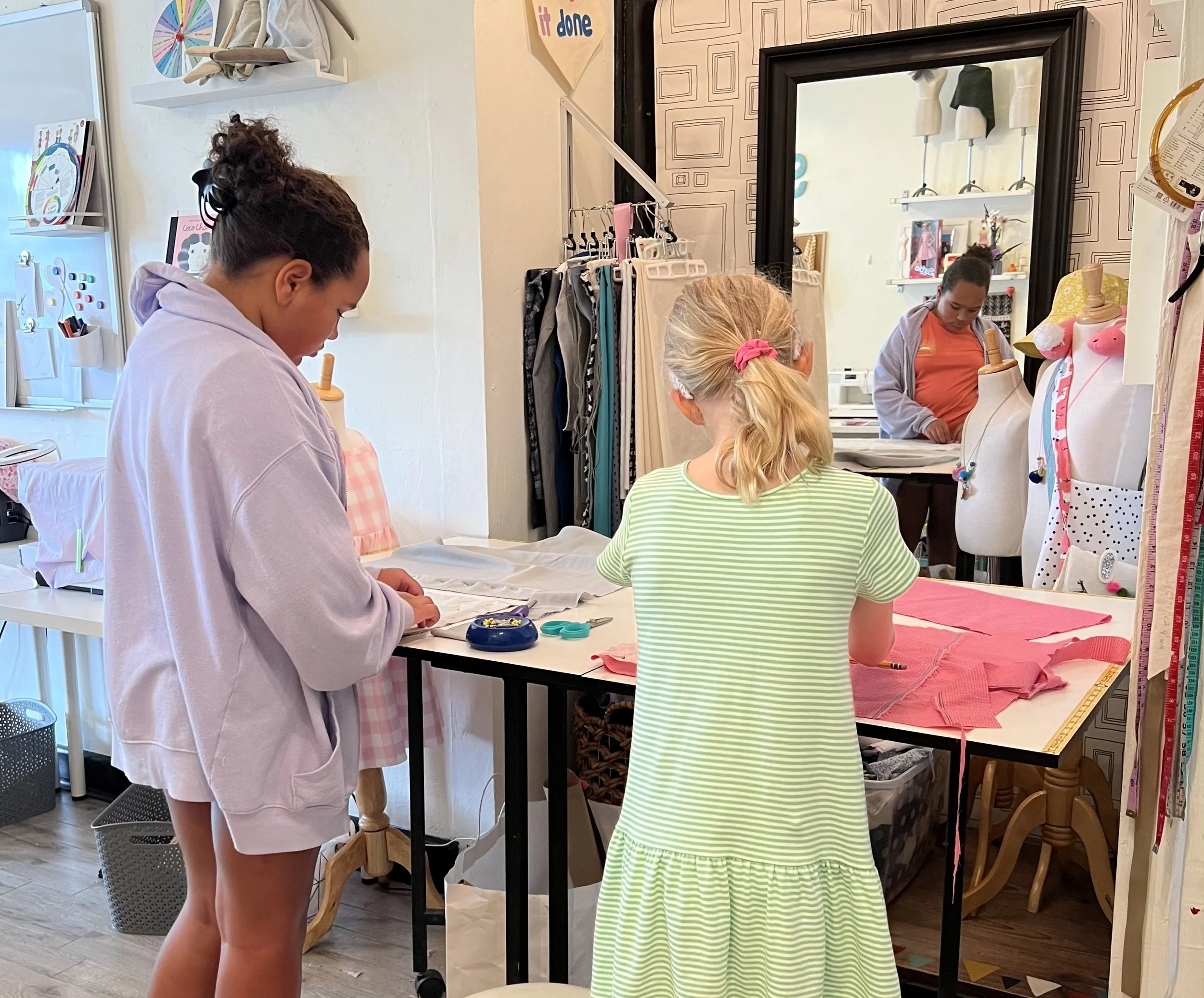 Three women working at a sewing or tailoring station inside a boutique or craft room, with fabric, scissors, and measuring tools.