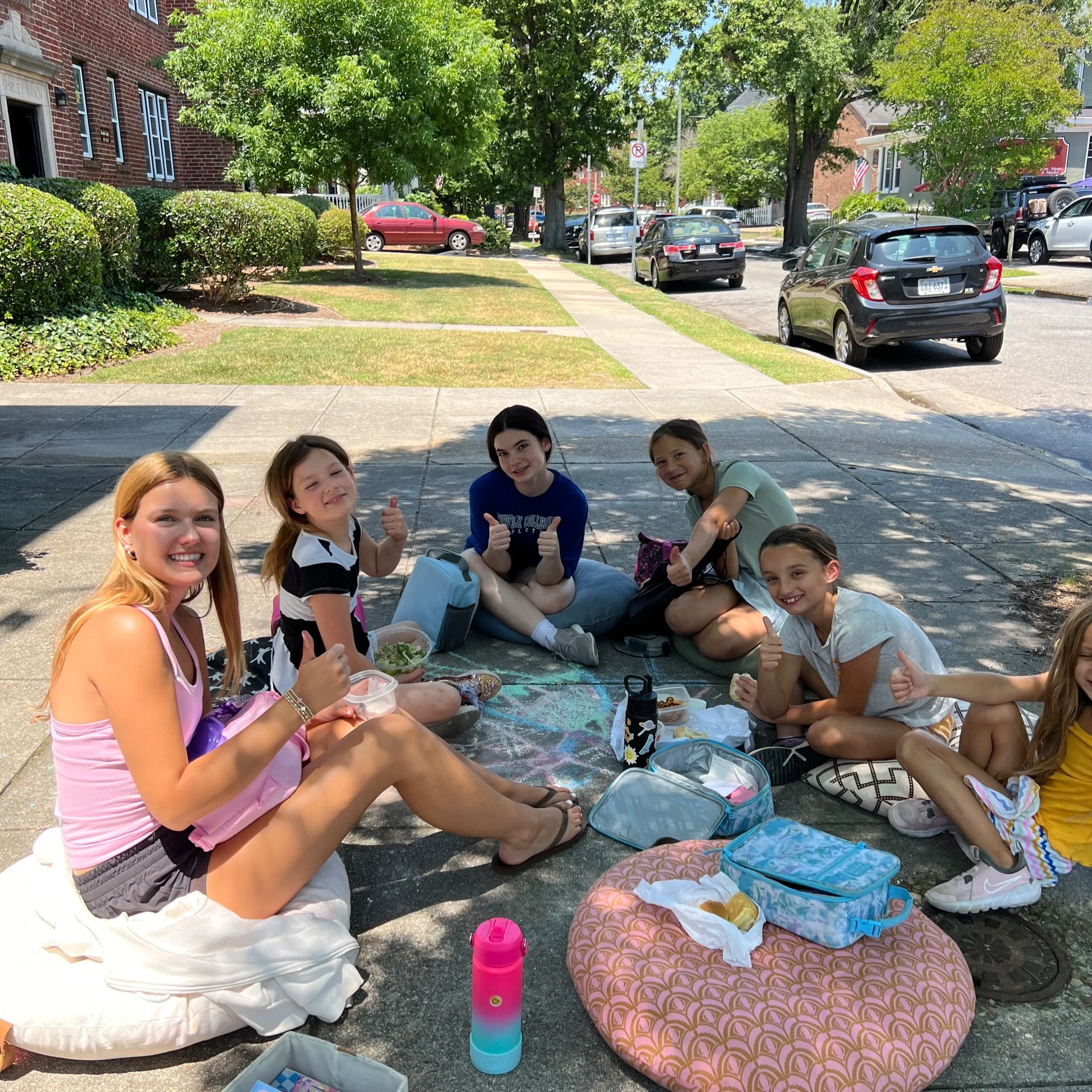 A group of seven children and a young woman sitting on a blanket and cushions during a picnic on a shaded sidewalk, smiling and giving thumbs up, with parked cars, trees, and houses in the background.