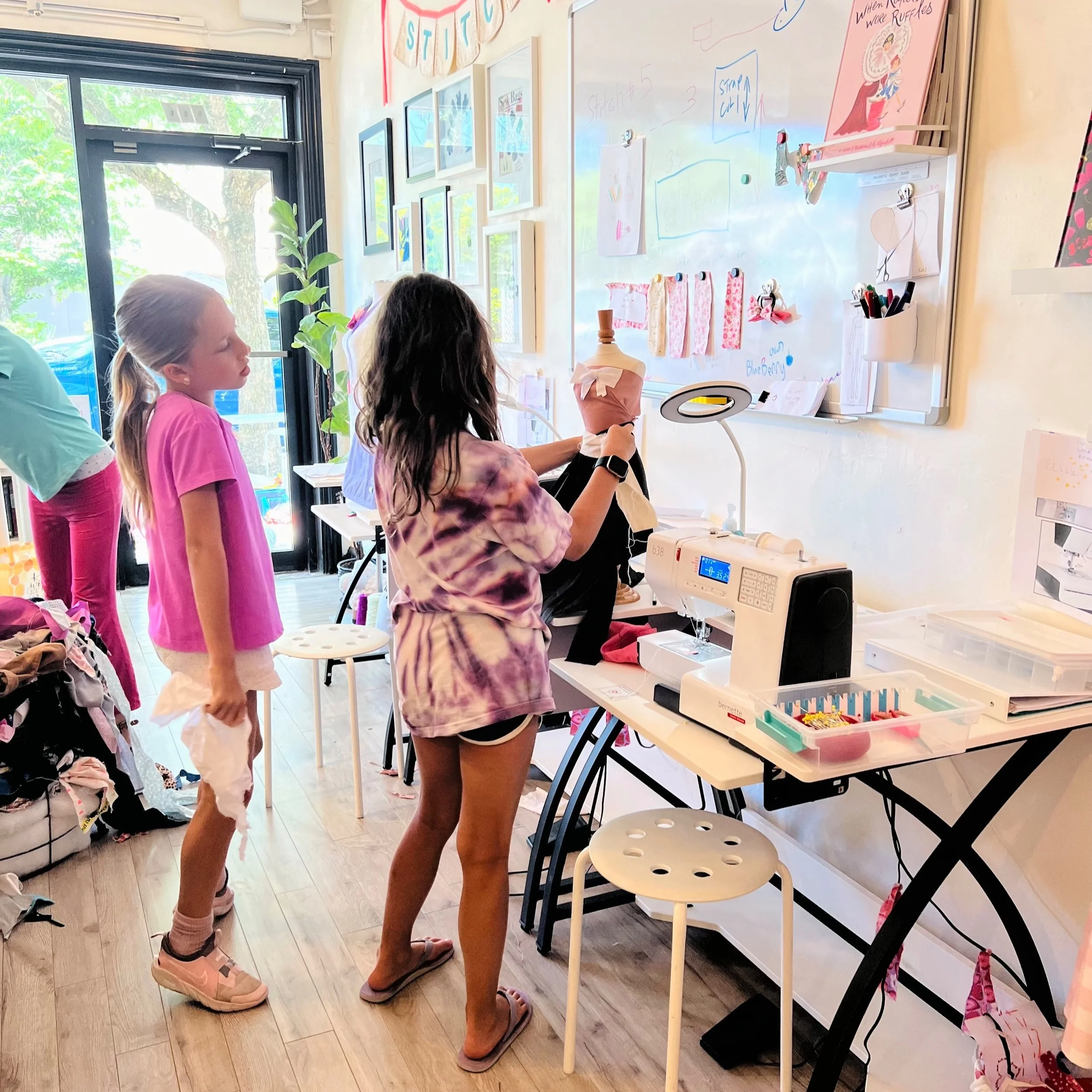 Three young girls in a craft room, with one working on a sewing project at a sewing machine while the others look on. The room has a whiteboard with drawings, colorful fabric swatches, and framed pictures on the wall. A glass door lets in natural lig