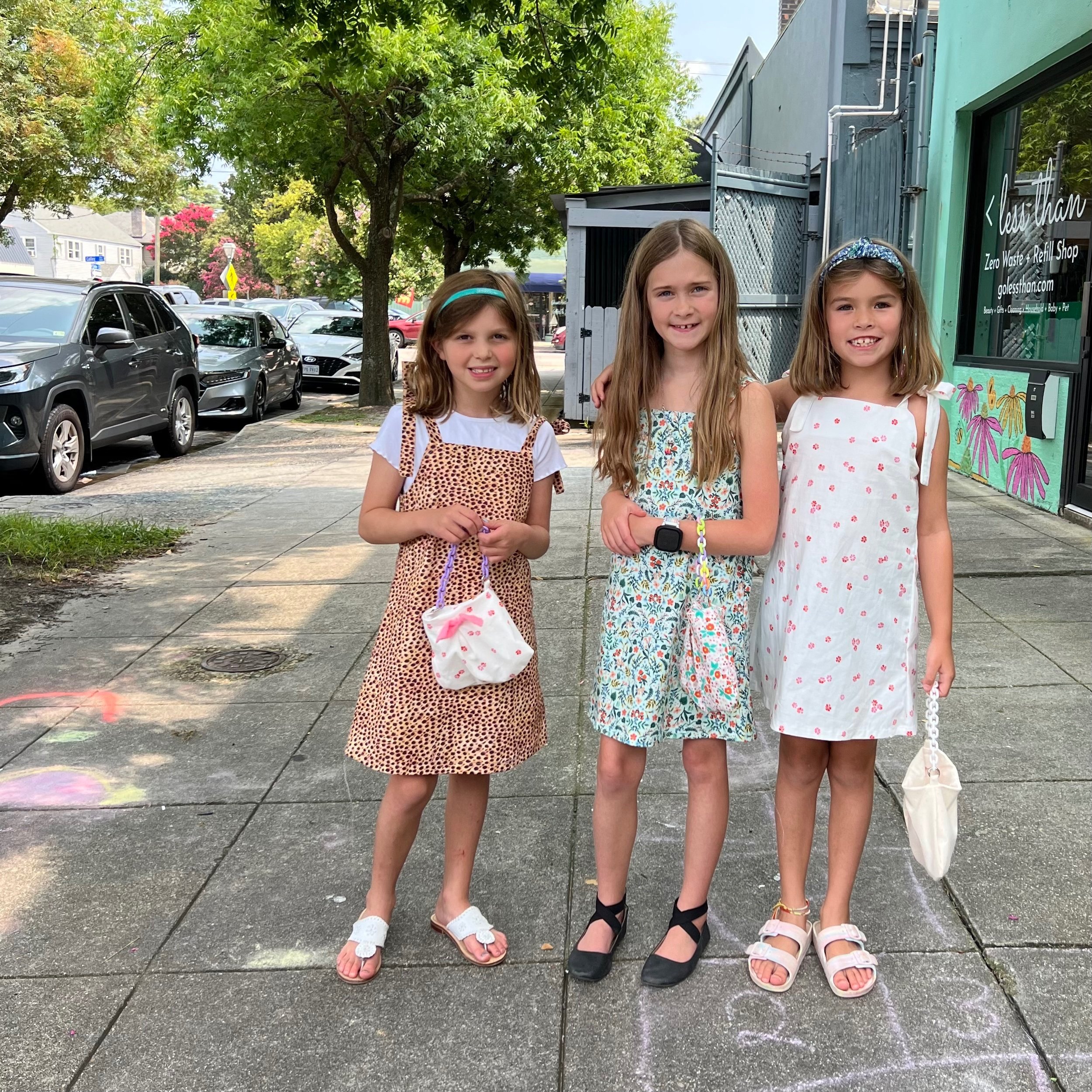 Three young girls standing on a sidewalk in a city, smiling and holding small bags. They are dressed in light summer dresses and sandals, with trees and parked cars in the background.