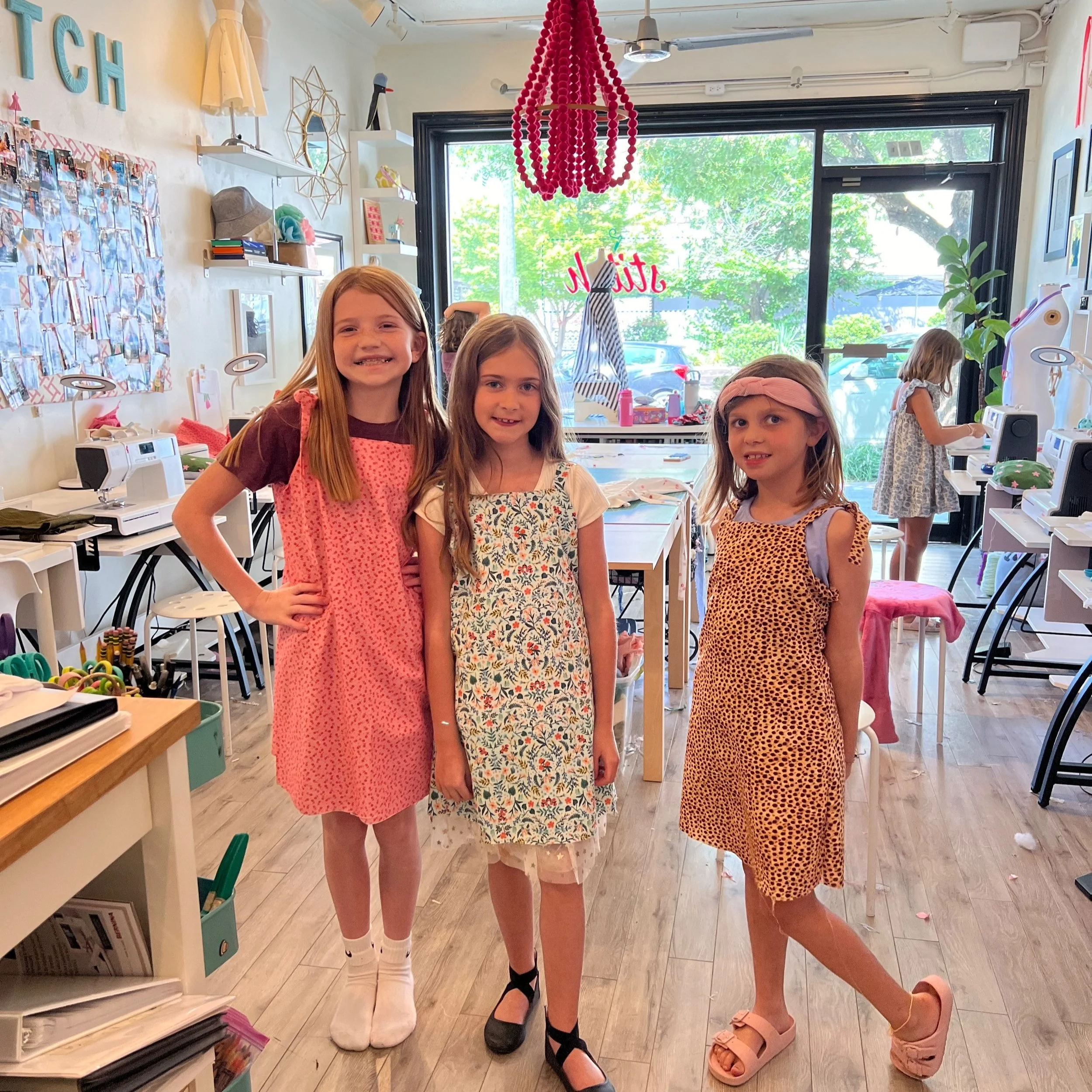 Three young girls standing inside a colorful sewing or crafting classroom, smiling at the camera. Two girls are wearing dresses and one has on a patterned dress with a pink headband. In the background, other children are engaged in activities at sewi