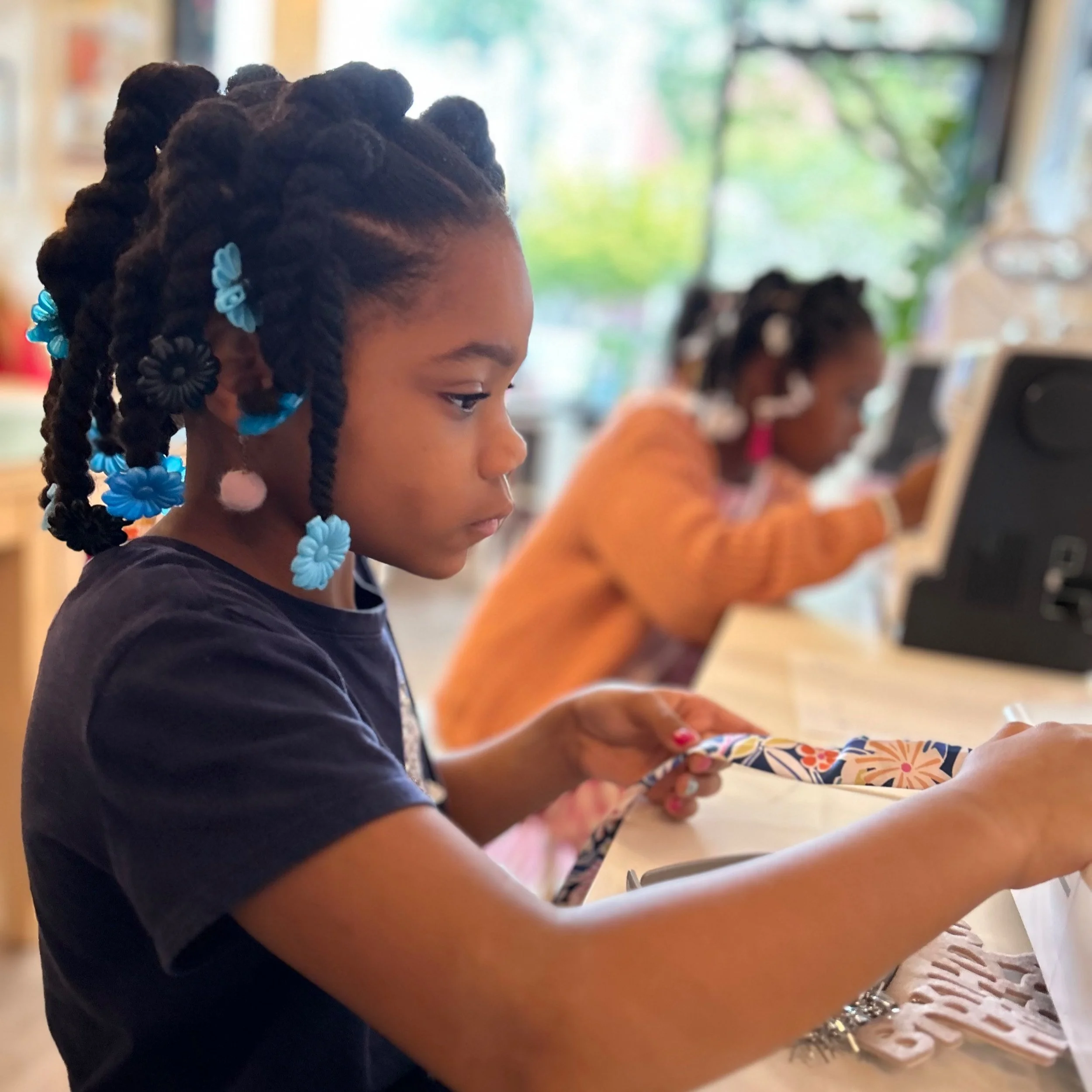 A young girl with braided hair adorned with blue and black flower beads is focused on a craft project at a table in a classroom or craft area, with another girl working on a similar activity in the background.