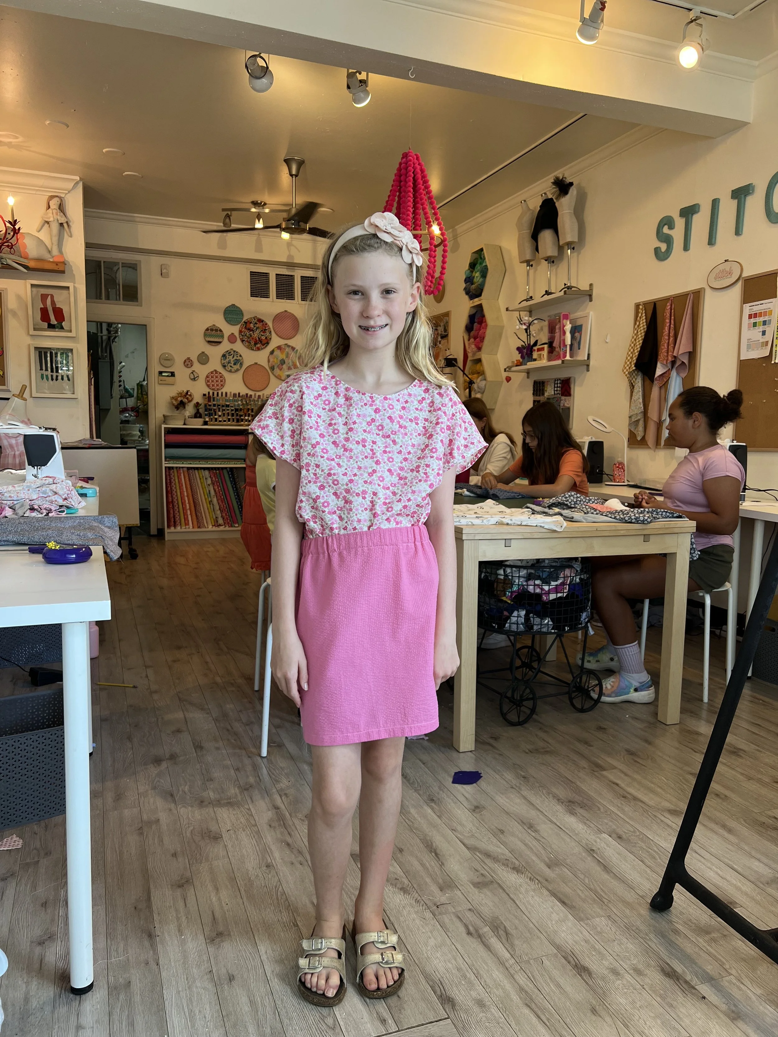 A young girl with blonde hair, wearing a pink floral top, pink skirt, and sandals, stands in a craft or sewing workshop, smiling at the camera. In the background, there are women working at a table, shelves with fabric, and decorative wall art.