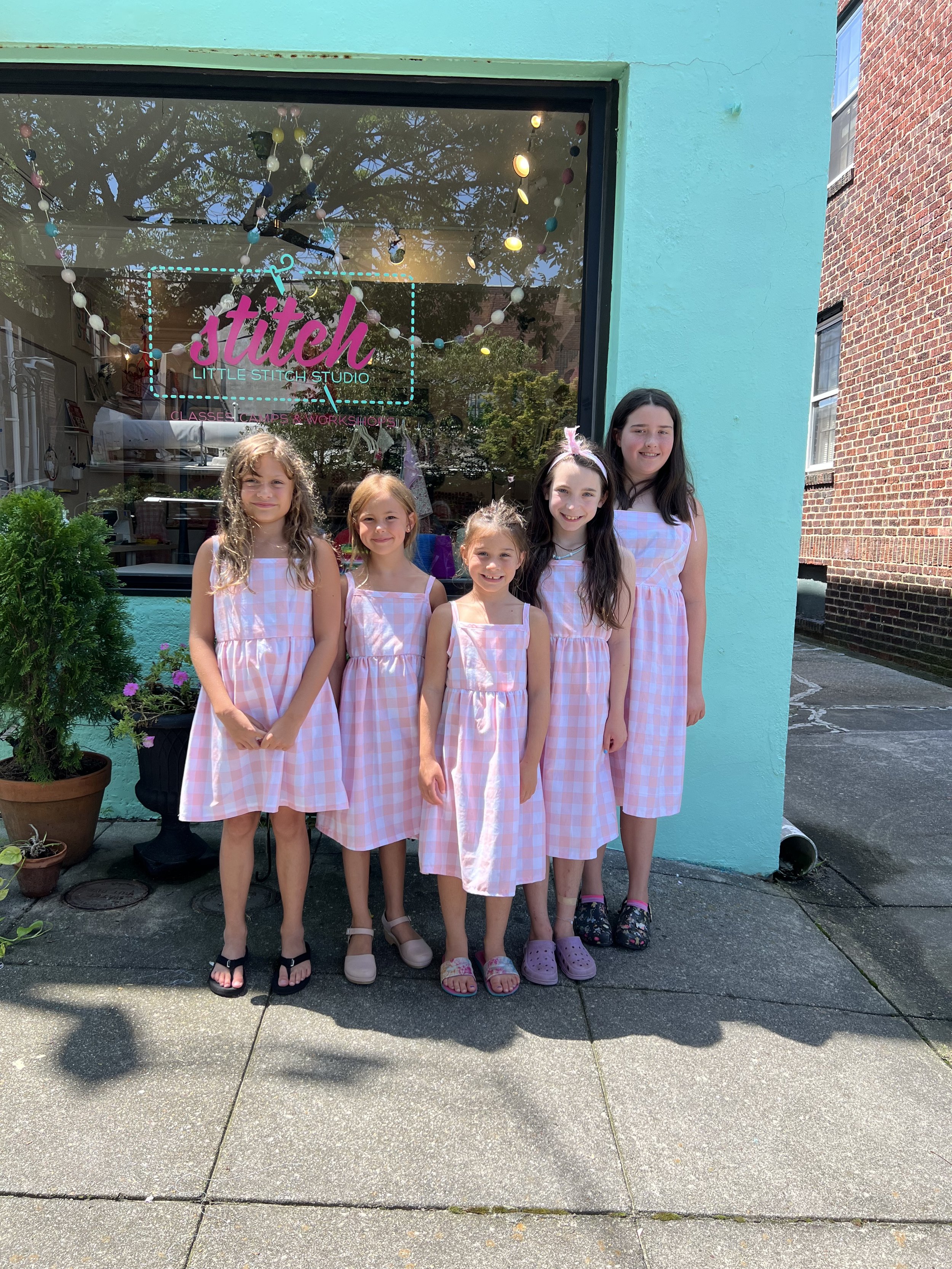 Five young girls standing outside a colorful storefront wearing pink and white checkered dresses. The window behind shows a sign that reads 'Stitch Little Stitch Studio' and decorations inside.