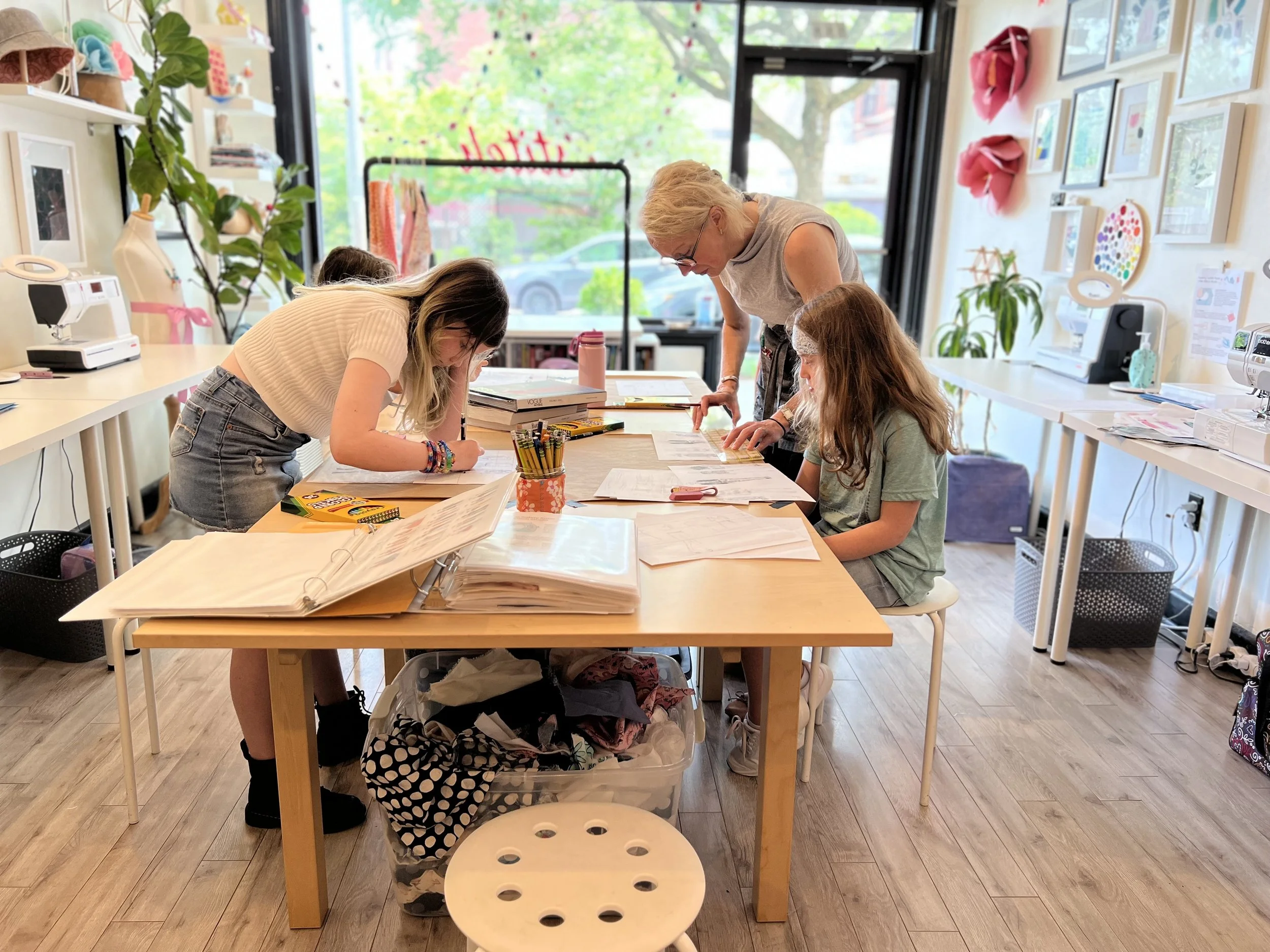 A teacher and three students working on assignments at a table in a brightly decorated classroom with art on the walls and large windows.