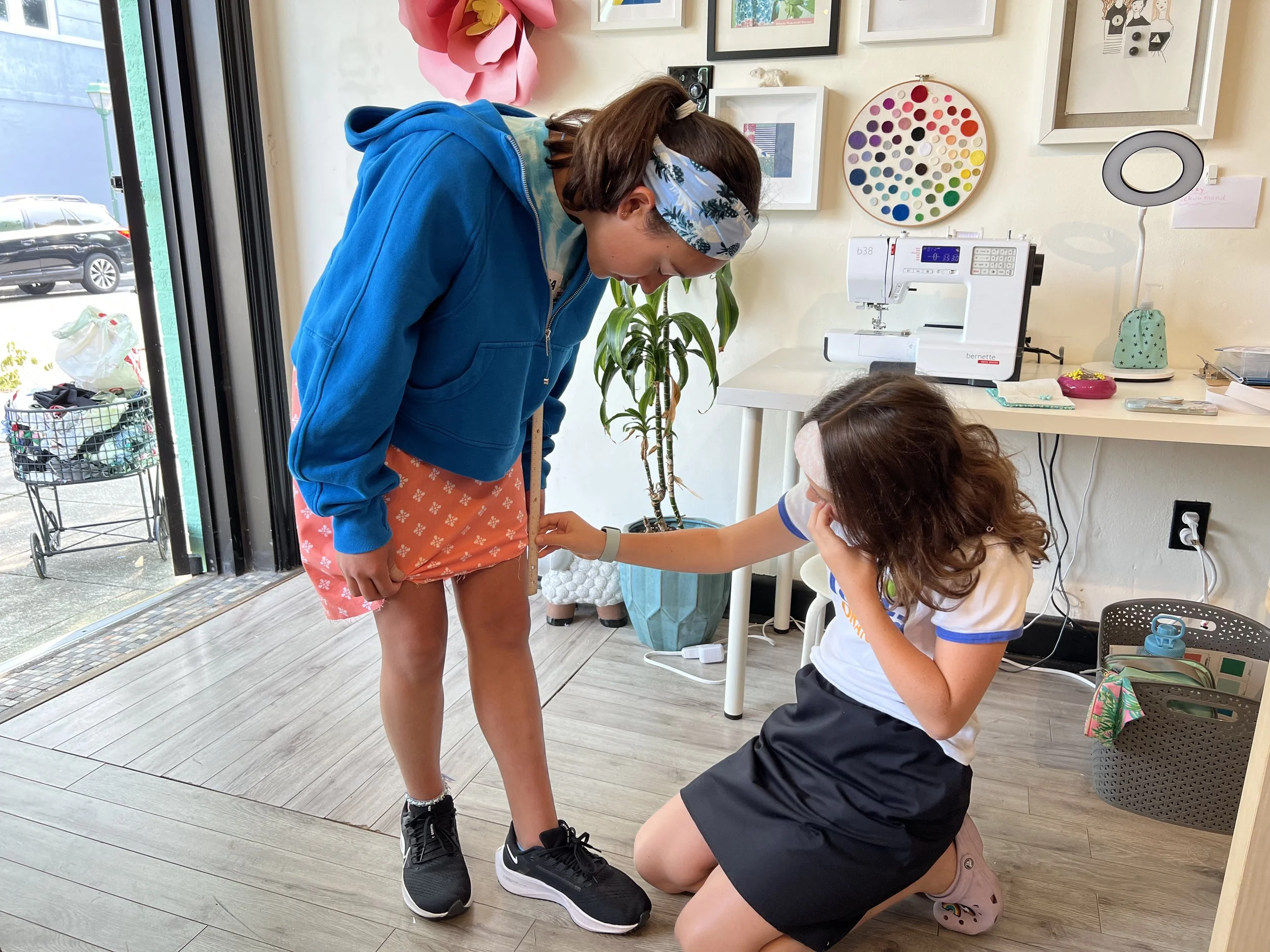 Two young girls are in a room, one standing with her head bent, the other kneeling and measuring her waist with a ruler. The room has a sewing machine on a white table, artwork on the wall, and a potted plant.