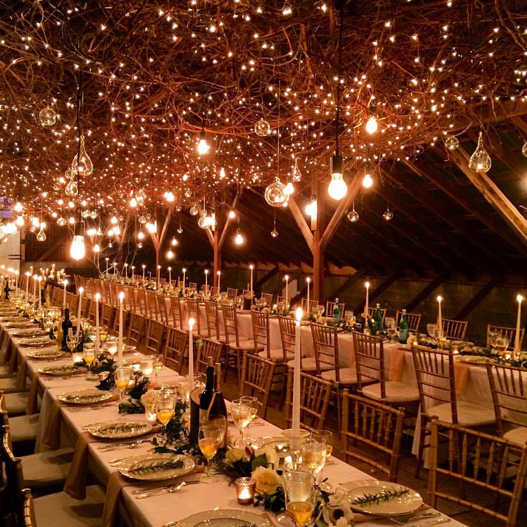 A beautiful lighted barn ceiling with twinkle lights, Edison bulbs, hanging candles and beautiful tables
