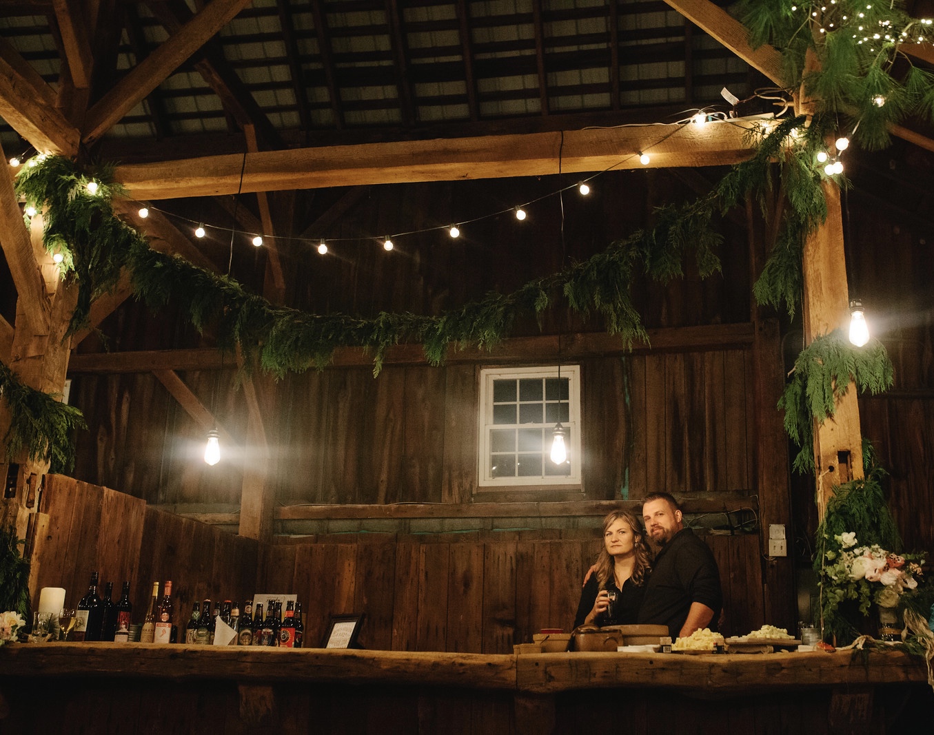 A super cool couple bartending at a winter barn reception with hanging greens