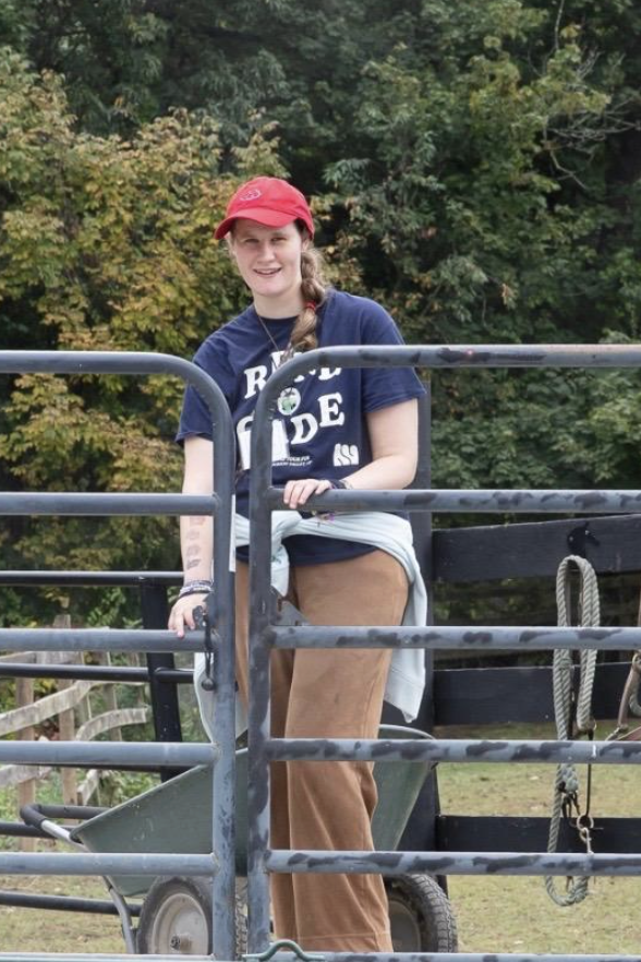A young woman standing on a flatbed trailer, wearing a red cap and a navy blue T-shirt, with trees in the background.