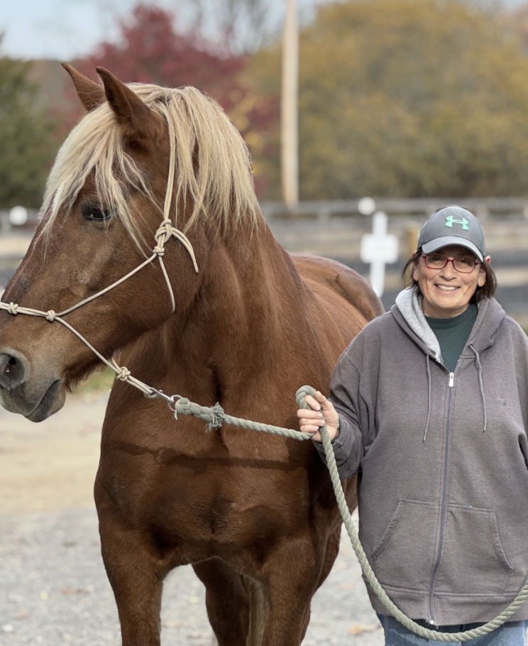 A smiling woman with glasses and a cap standing outdoors next to a brown horse with a blonde mane, holding its lead rope. The background features autumn trees.