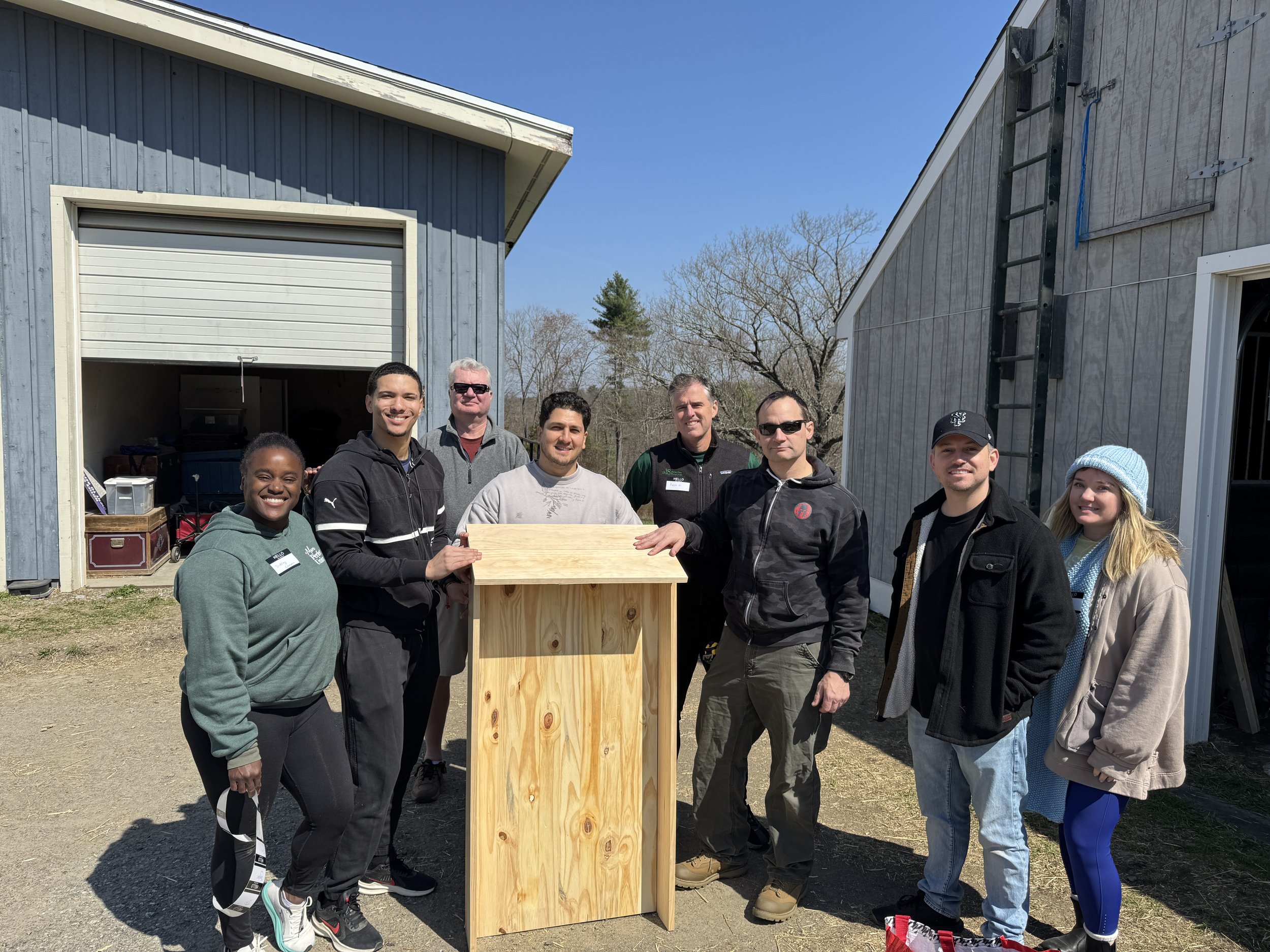 Group of eight people standing outdoors around a wooden structure, with blue sky and trees in the background, some people wearing sunglasses and others smiling.