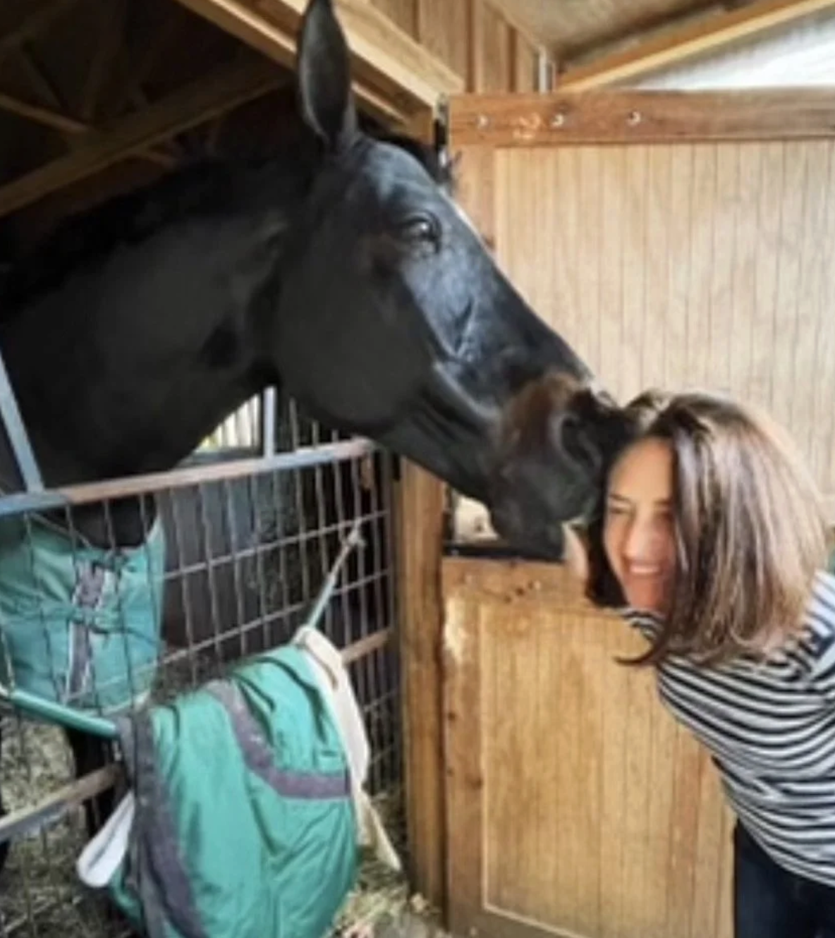 A woman with shoulder-length brown hair and a striped shirt is smiling as a large black horse leans its head over a stall door to nuzzle her affectionately in a stable.