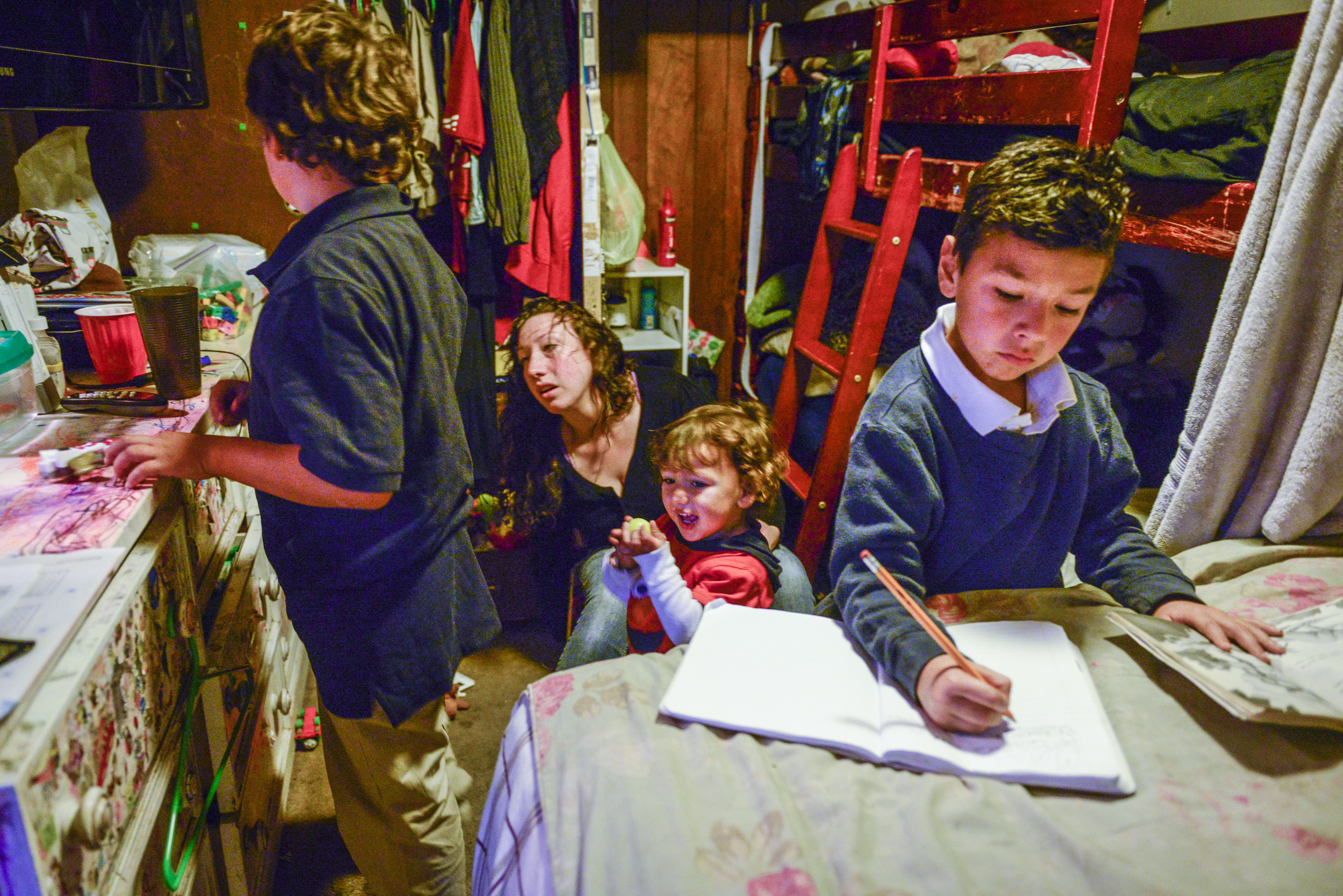 Mateo, 7, finishes his homework while Amanda helps Dominic and Christopher get ready to go to school. The studio they live in is so small there is no room for a table. The kids do their homework on their bed.