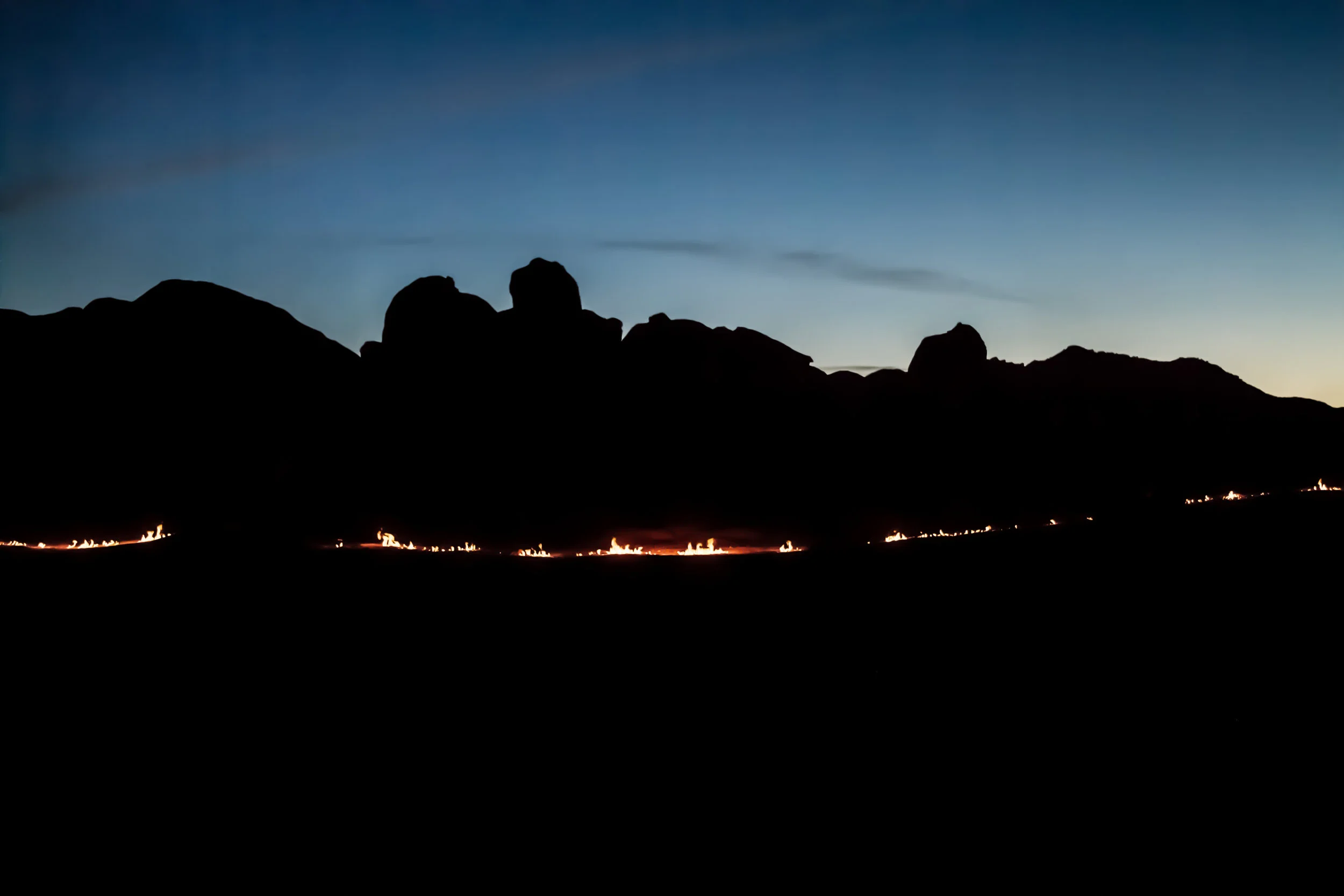 Horizon Parallel 2015Flame, Motorcycles 70' x 10" x 10" Featured in La Motocyclette. The Mojave Desert is a place the American psyche has turned it’s heart to, a unique place in our personal mythology. The poetry of the harshest and most rugged terrain upon which some of our go west, rugged individualism, survivalist identity has been formed. The vastness, the quiet, the trackless landscape. The playas, dried remnant of a prehistoric time are symbol to the ever changing, and the slow timescale of the earth. There is an open story of abstraction and projection alongside the prehistoric remnants that makeup the Mojave. This famous California desert has the perfect intersection between off-road culture, speed trials and earth art, both in it’s history and also it’s present. I was inspired, and I wanted to work on a sculpture that dialogued with this mythopoetic space. It only seemed fitting to create this piece out in the backcountry, taking a journey and setting out to see if I could find one of those iconic vast Mojave canvases that was that was blank, unpaved, off road and not accessible by anything but bike. This place existed in my mind, but I had to go find it. Riding out into backcountry is tough. You aren’t really going fast. You’re in soft sand and picking your way through it. We were riding vintage enduros back there. And one of the bikes was from ’74. These bikes are different, they are heavier and they don’t handle like a modern dirtbike. But that’s the way it is out in the wild. Everything depends on you, having enough water, knowing how to read the weather, how to backtrack and know your orientation, how to stay warm, make your own light and so on. That is survival, you really have to learn to depend on yourself, but that feeling of independence, and ultimately interdependence with the earth, is really in my opinion, one of our most fundamental powers and a very real source of joy. We were all packed up over the back of the bikes. And that’s why I love those bikes - they are tough and well built and you can take them anywhere. The whole world is there for you to roam and in the desert - it’s pretty wide open - so you can ride anywhere. We were picking over rocks and into new territory. We were riding on backroads and dried lake beds. We spied this small passageway up and through the rocks that we could slip through. Just through this rock crevice canyon was this phenomenal natural amphitheater, bowl shaped enclave, hidden. It was dusk, so we unpacked to the sounds of the coyotes down in the playa below. Most of my art practice is based on getting us back in touch with some latent instincts that I think we left out there in the bush. While we were riding during night fall, we really got that sensation of the wide open blank vast space, the giant infinity around us, and the open sky. That’s not an intellectual experience, its a sensual one. No matter which direction, you are riding toward a horizon, something far and ahead of you that you can’t see. You are making a perpendicular axis, dividing the circular earth with parallel lines, dividing the circle boundary of your range of vision. You sort of spread out over the land spatially. You start to think of yourself separating yesterday and tomorrow and there’s a relationship to speed. It felt as though we were out there in the middle of nowhere cutting the day and night in half. Human beings are a gold mine. Boldness, backcountry, riding, sensual experience that goes out beyond your skin, loudness, speed, these aren’t just concepts, these are lifestyles. Sometimes you have to burn a blaze, or head out into the middle of nowhere, or ride desert at night, to enliven your sensuality. You are a shapeshifter and you’ve got the power. It’s ancient, it’s in your skin.