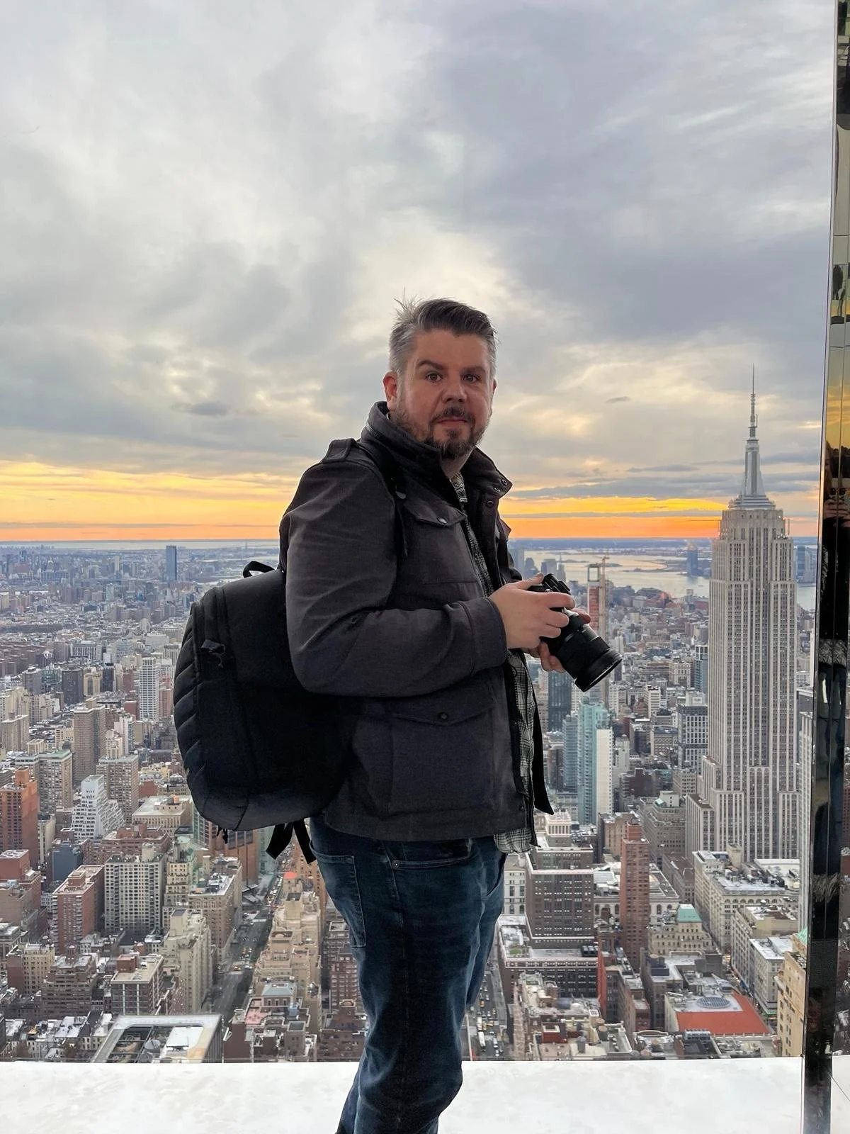 A man with a camera in front of the New York City skyline, including the Empire State Building and the Hudson River at sunset.