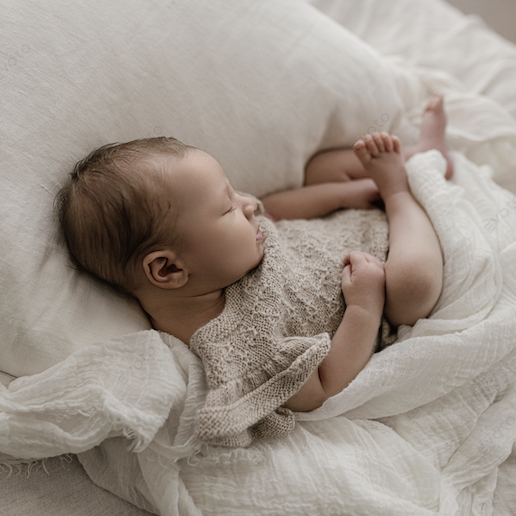 A sleeping baby in a beige knitted outfit lying on a cream-colored bed with white pillows and blanket.