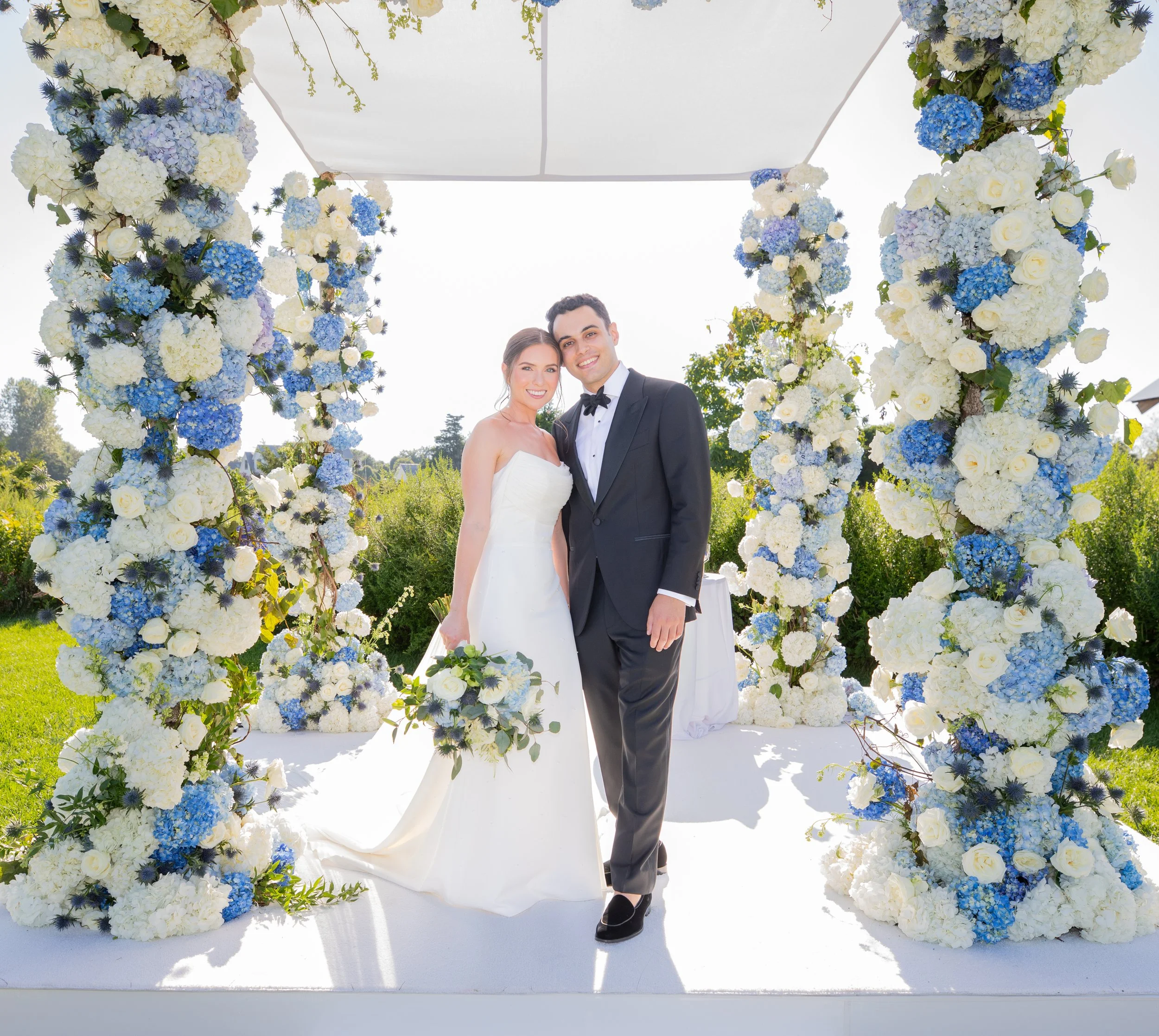 Luxury outdoor wedding ceremony in the Hamptons with bride and groom under blue and white floral arch.