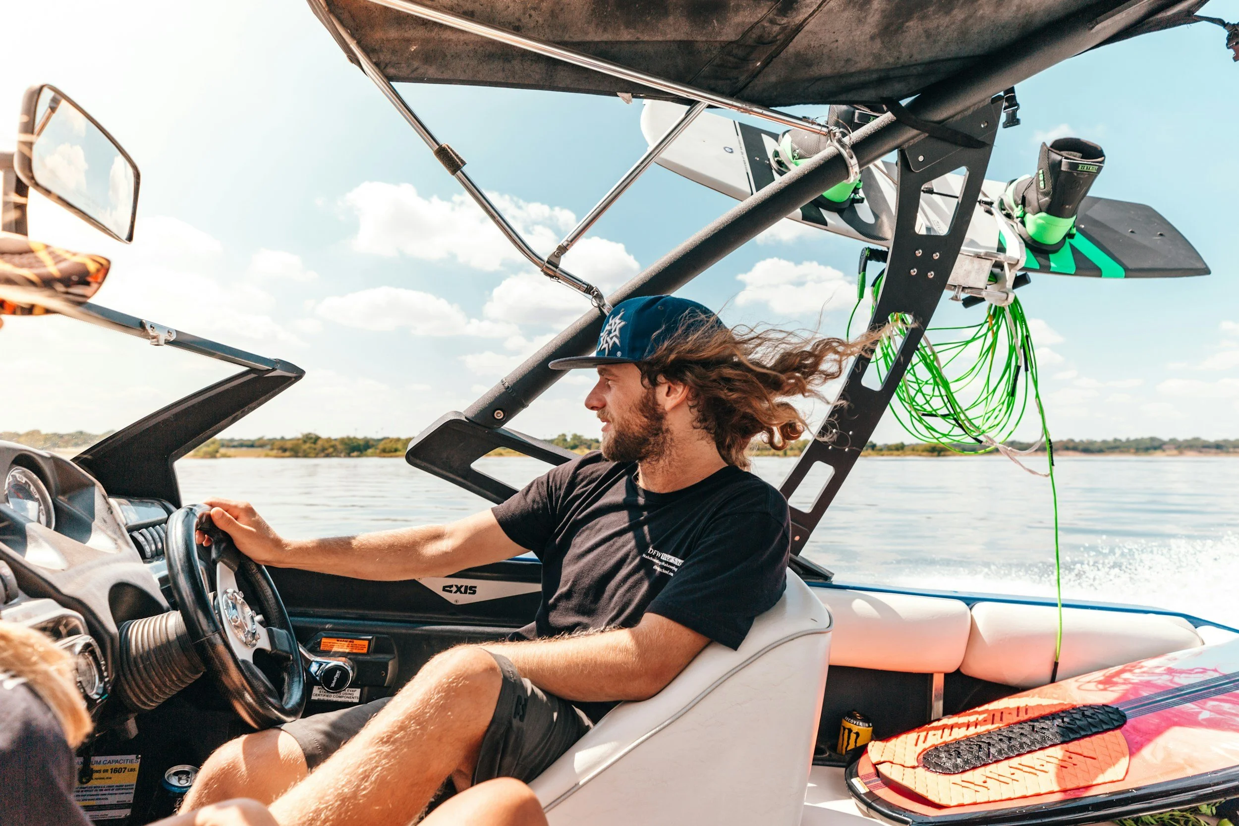 A man with long, flowing hair and wearing a blue baseball cap and black T-shirt driving a motorboat on a lake with a clear sky and some clouds.