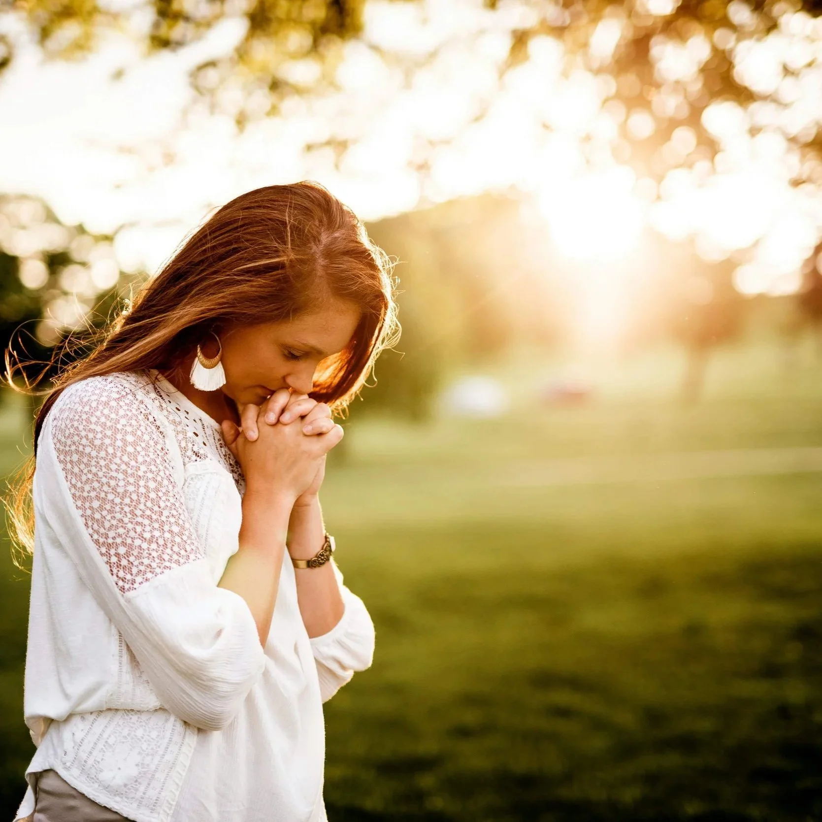Woman with clasped hands standing in grassy field with trees and sunlight behind her.