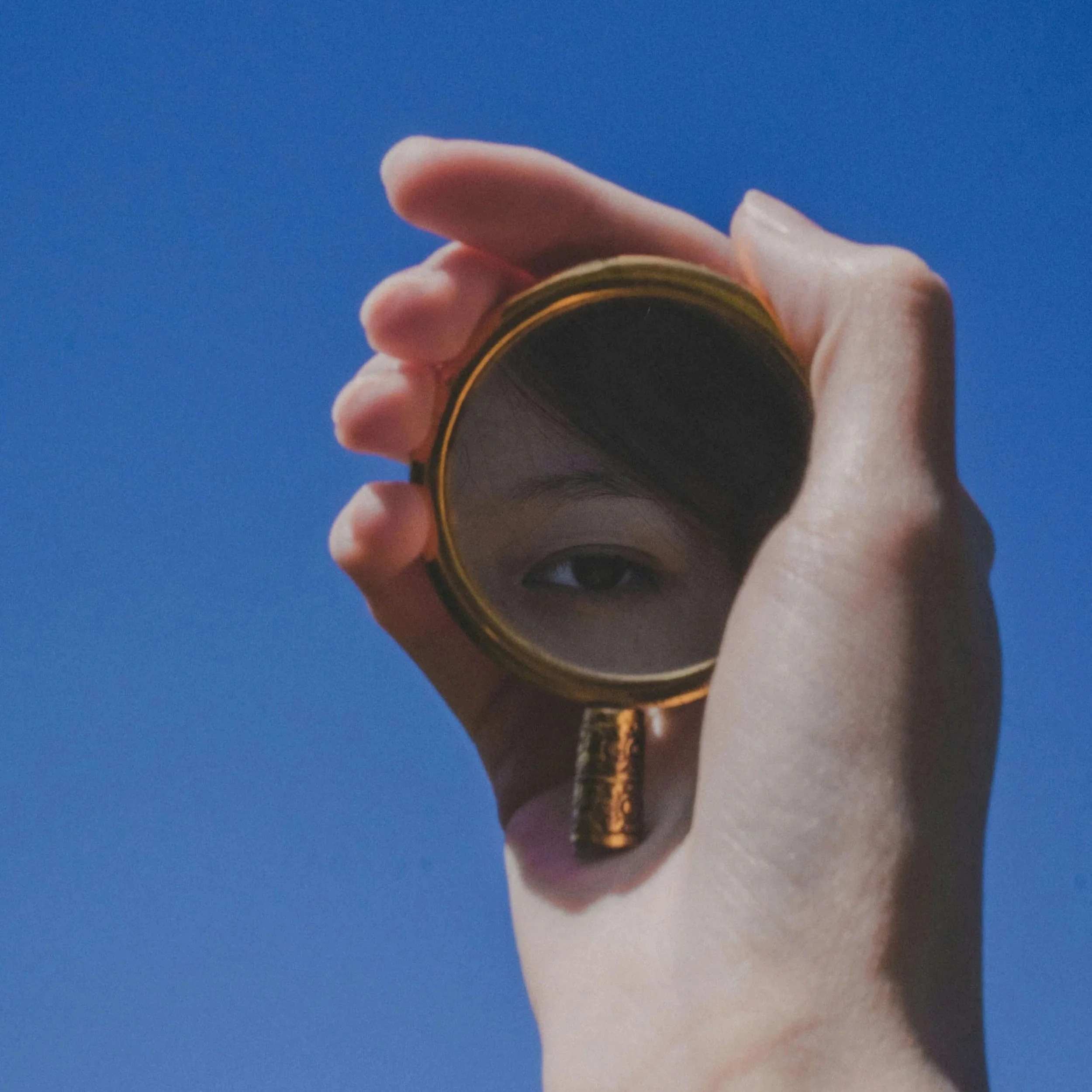 Hand holding a small mirror reflectins an eye with blue sky background.