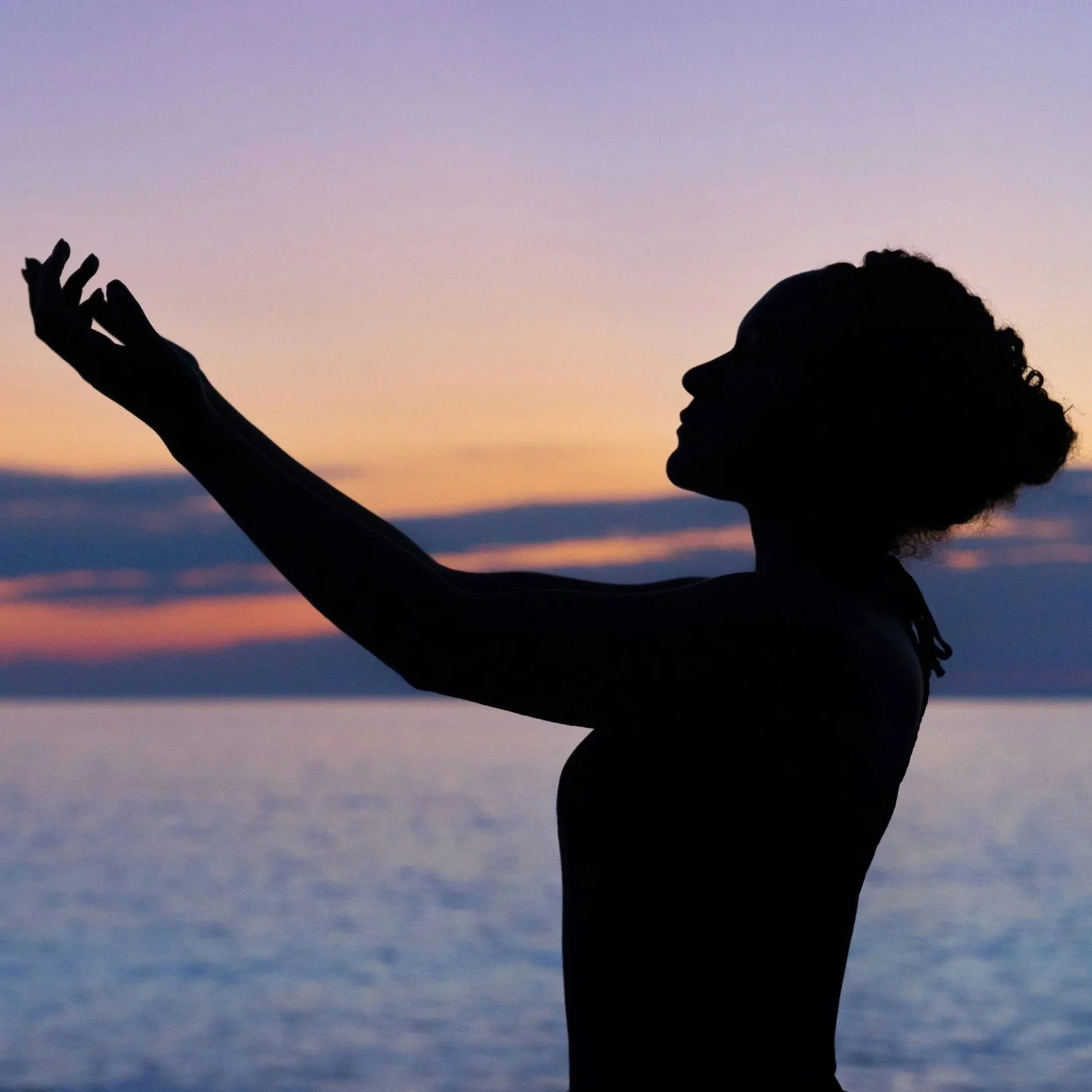 Silhouette of a woman standing sideways with outstretched arms in front of water at sunset.