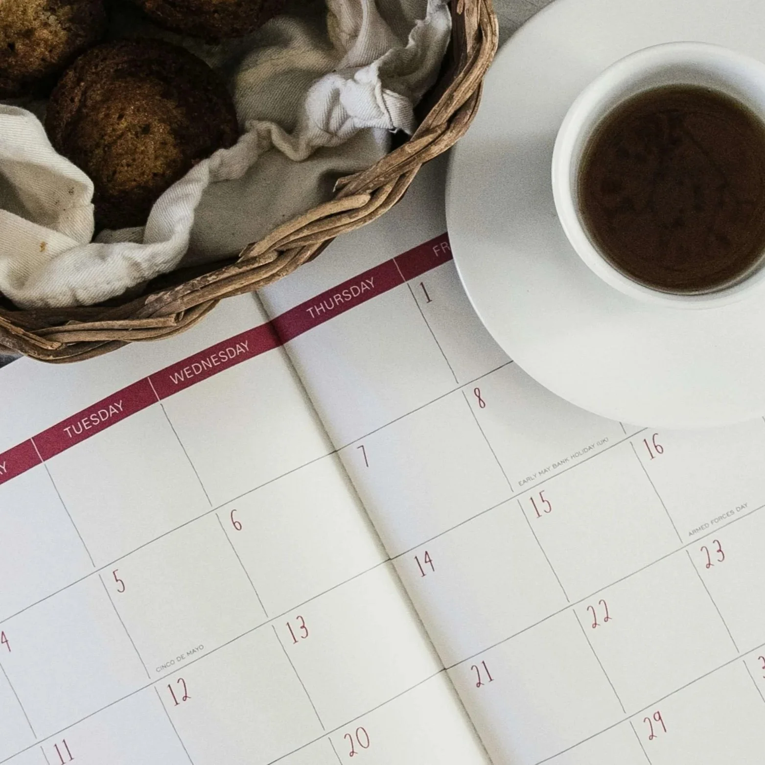 An open calendar with a cup of coffee and basket of muffins.