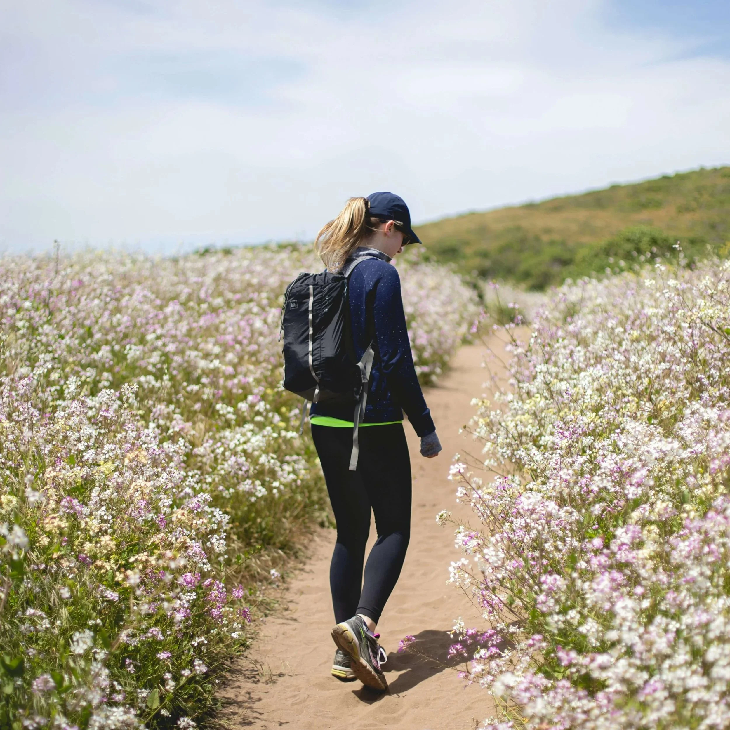 Woman walking on a dirt path looking at the wildflowers growing alongside it.