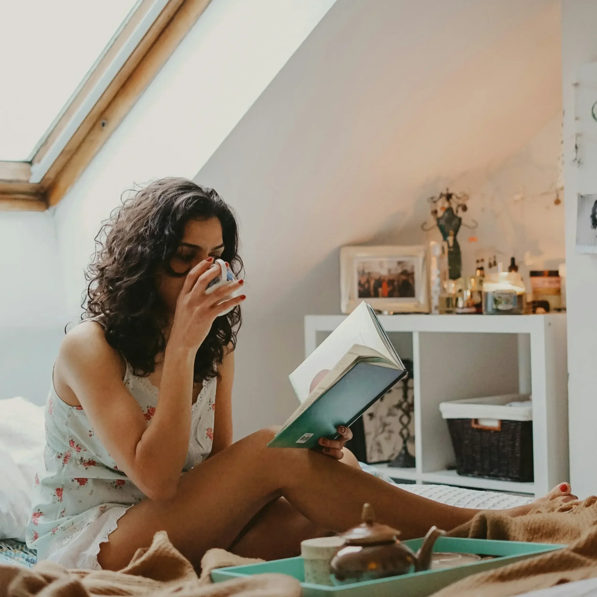 Woman relaxed in bed reading and sipping from cup.
