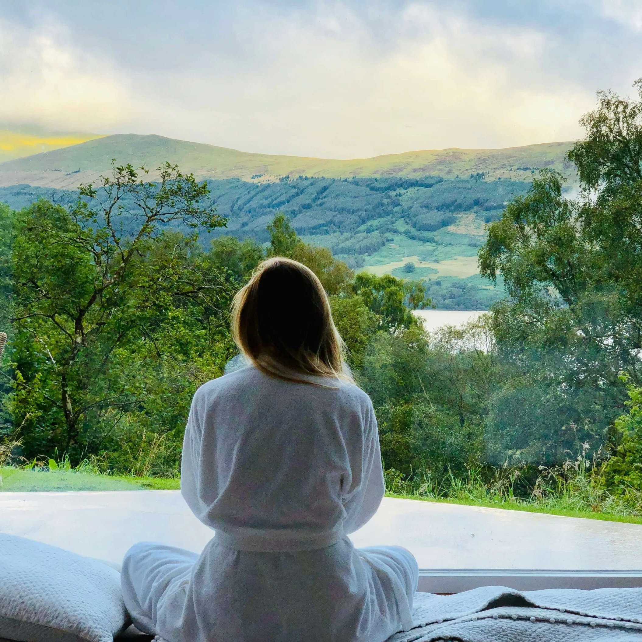 Woman in white robe sitting on deck overlooking trees and mountains.