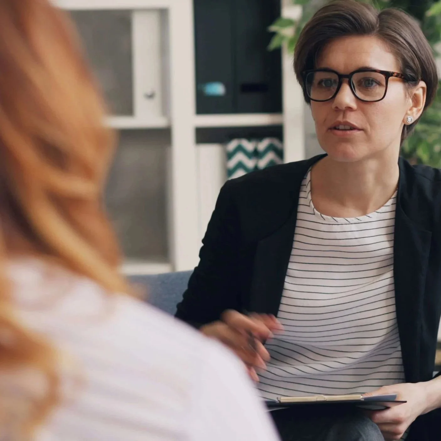 Dark-haired woman with glasses talking to another woman who has her back to us.
