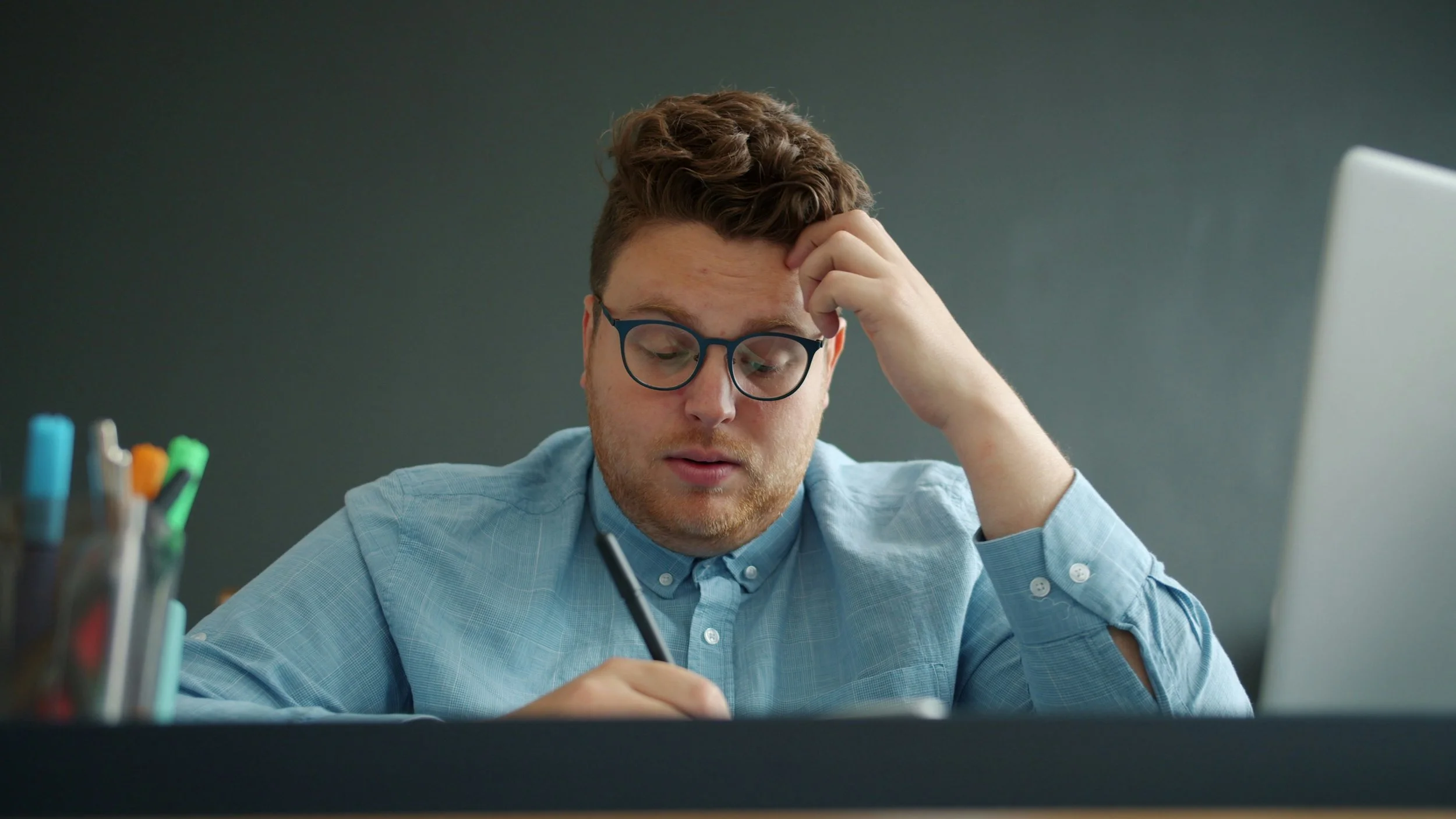 Male teacher sitting at desk holding a pen with his head resting against his hand looking stressed.