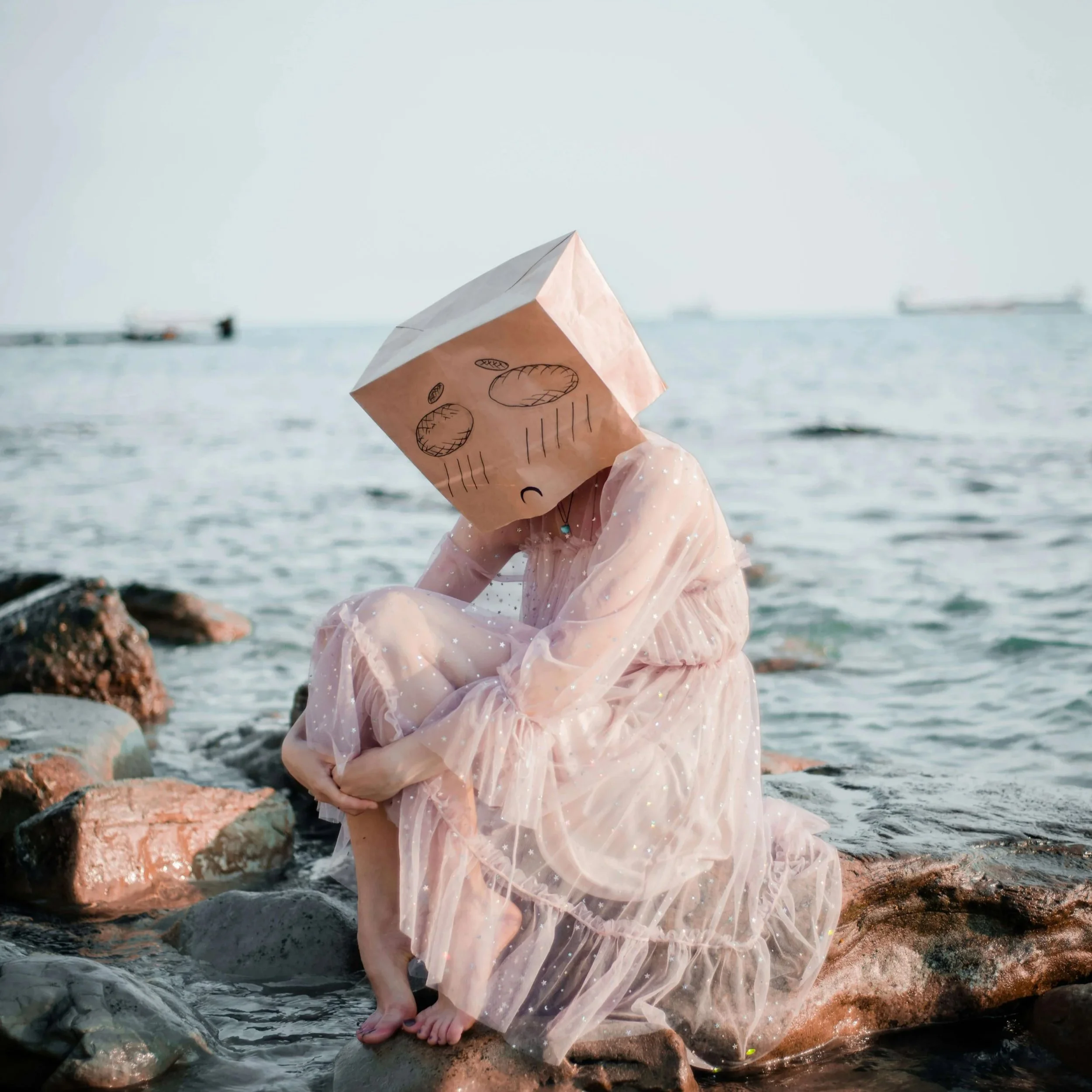 Woman in white dress sitting on a rock with a bag on her head that has a frown drawn on it.