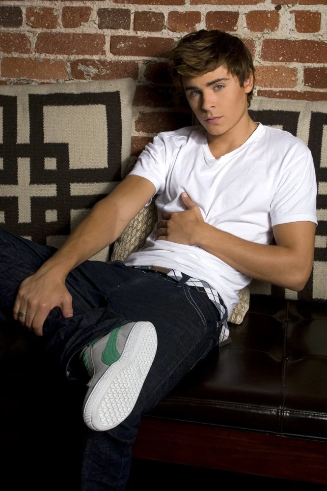 Young man with brown hair and light skin sitting on a black leather couch with a pillow, wearing a white t-shirt, black jeans, and gray sneakers with green accents, in front of a brick wall.