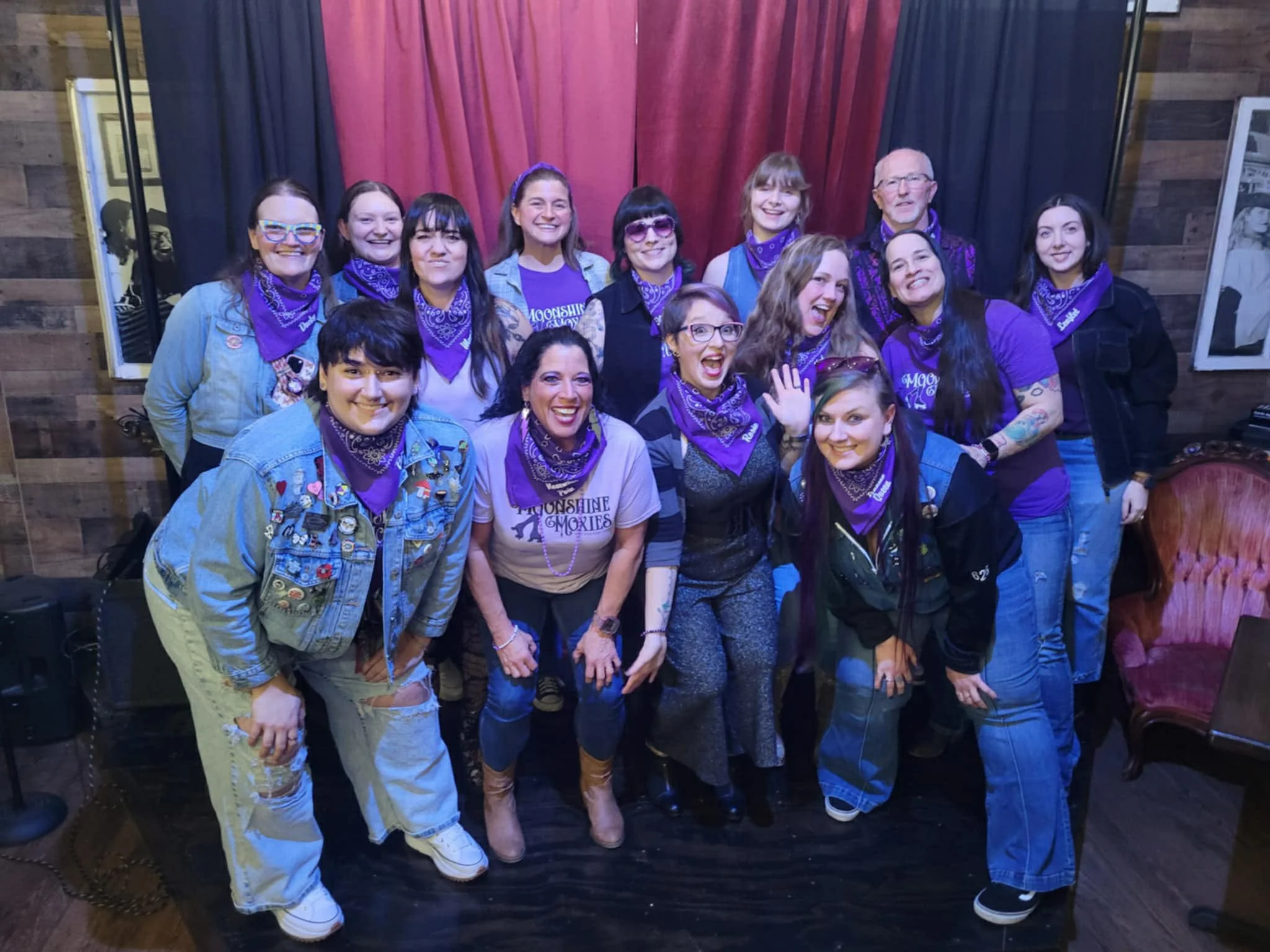 Group photo of the Moonshine Moxies at a draft party. They are all wearing a purple bandana and many are wearing other purple clothing. Panic is in the back row, smiling widely.
