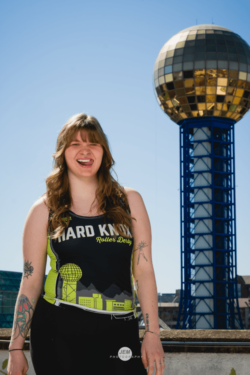 Headshot of Panic Attack. She is in her black HKRD uniform with the Sunsphere in the background. Her hip is cocked to the side, she is winking, and she had her tongue catching the side of her lip, all in a sassy way.
